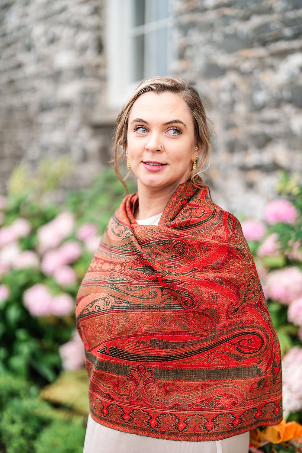 Woman wearing ornate red paisley shawl against stone wall with pink flowers.