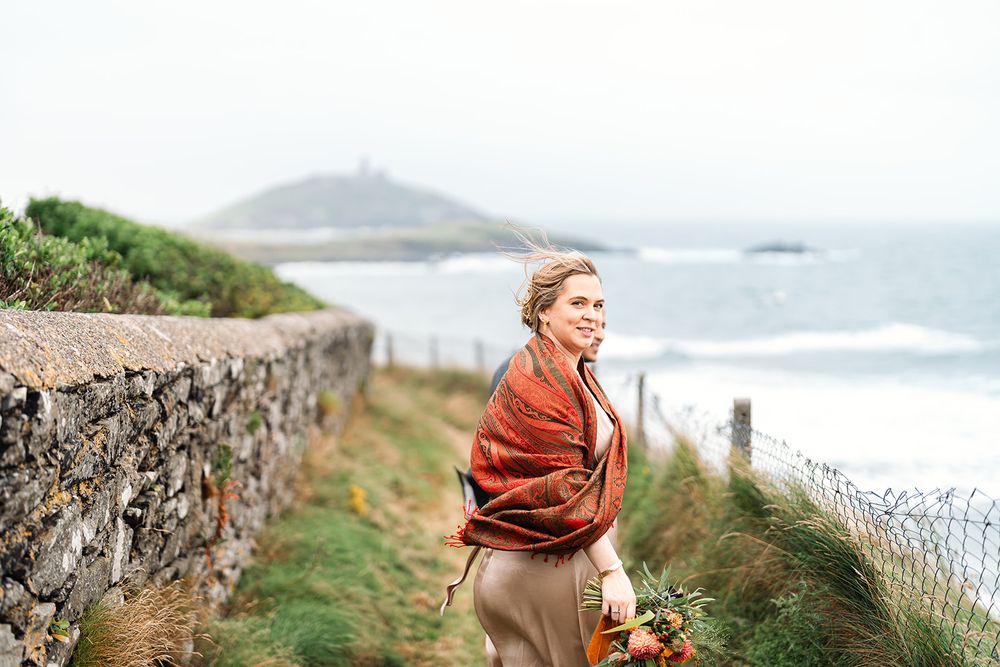 Figure in rust-colored wrap standing by coastal stone wall with flower bouquet.