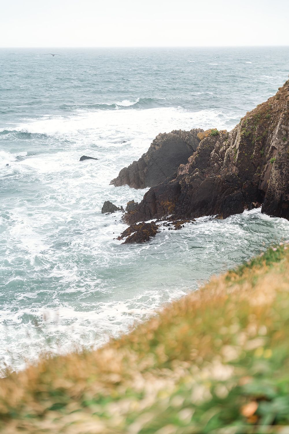 Rocky coastline with crashing waves against rugged cliffs and ocean spray on a misty day.