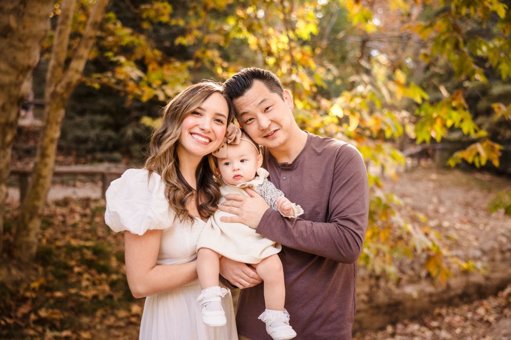 A family of three poses together outdoors during autumn with golden leaves in the background.