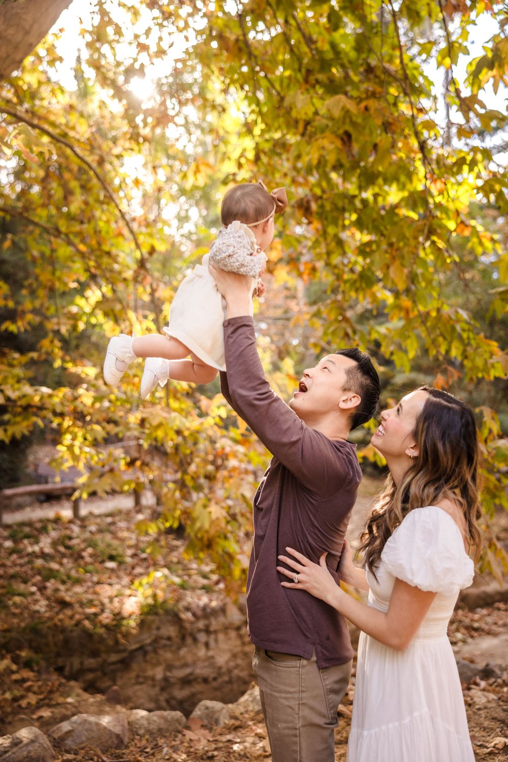 A couple lifts up a small child together while enjoying a sunny autumn day in nature with golden foliage in the background.