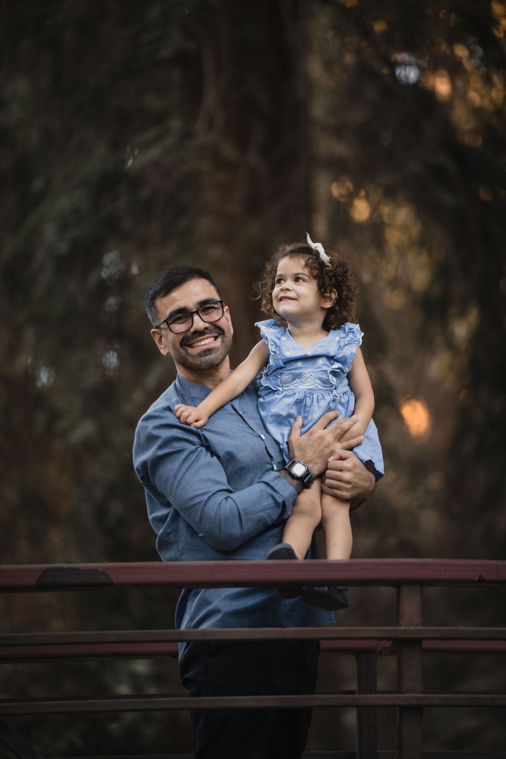 An adult and child share a sweet moment together on a wooden bridge in a wooded setting at dusk.