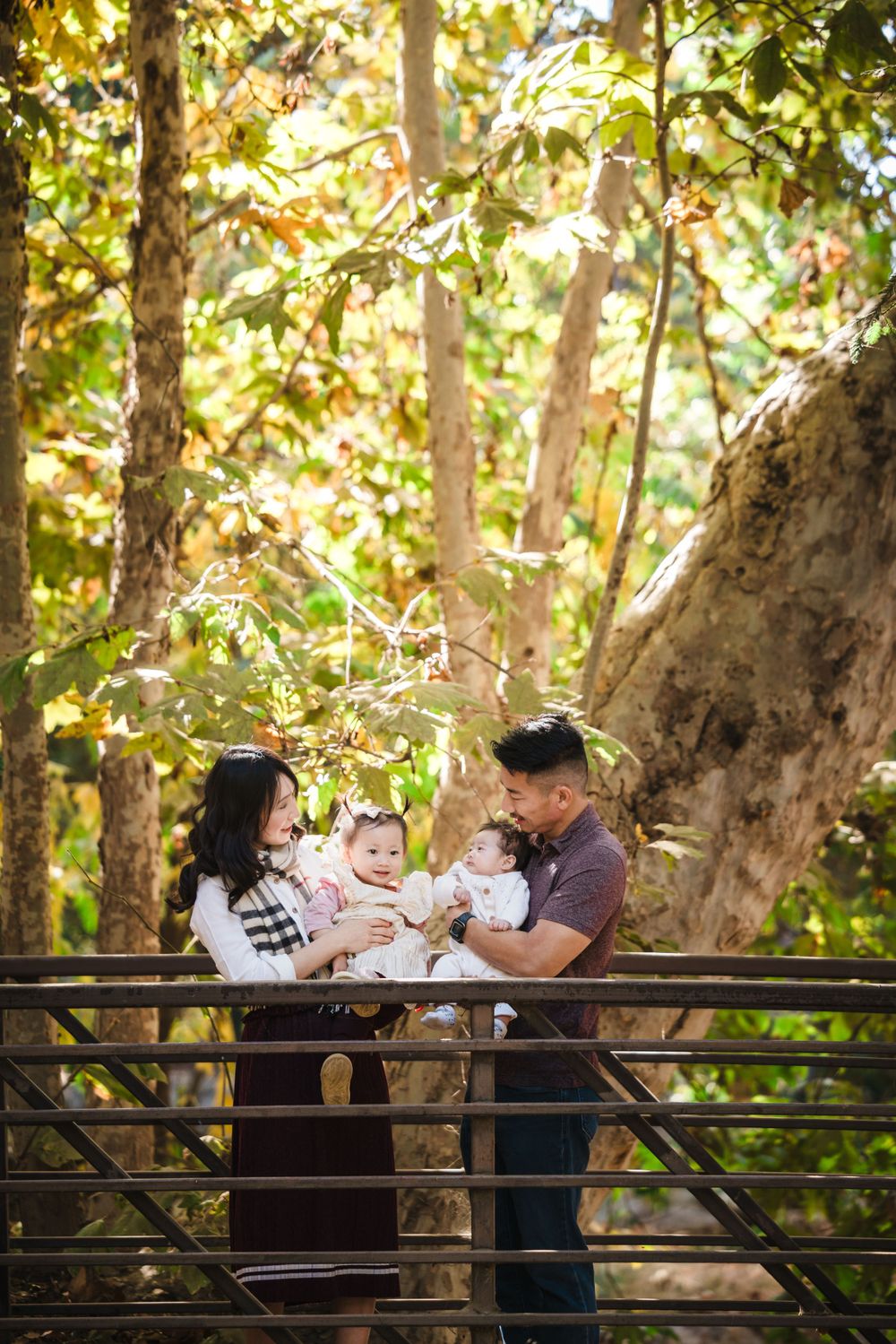 A couple stands on a wooden bridge in a sunlit forest park holding their baby.