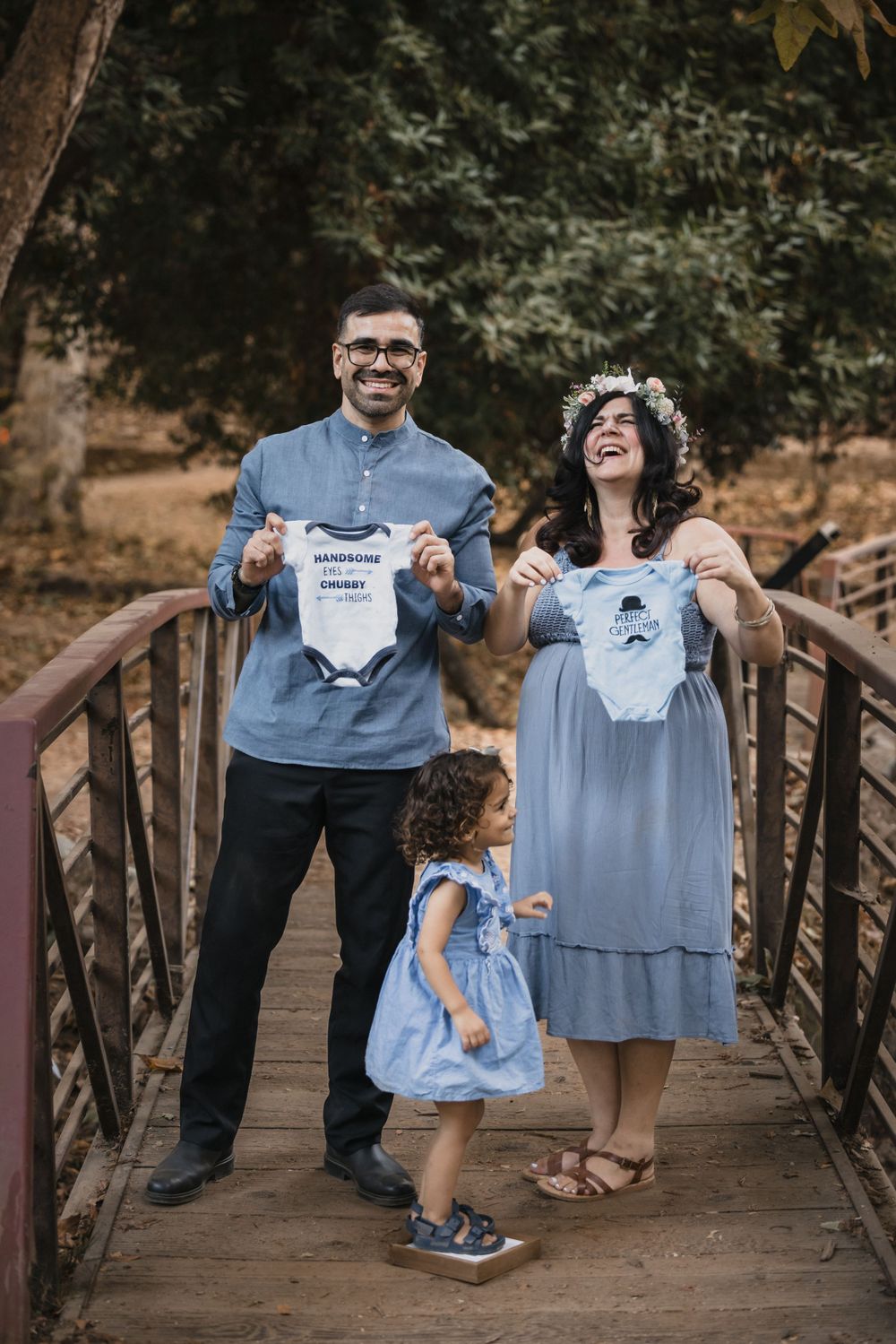 A couple and toddler stand on a wooden bridge holding baby onesies in a nature setting.