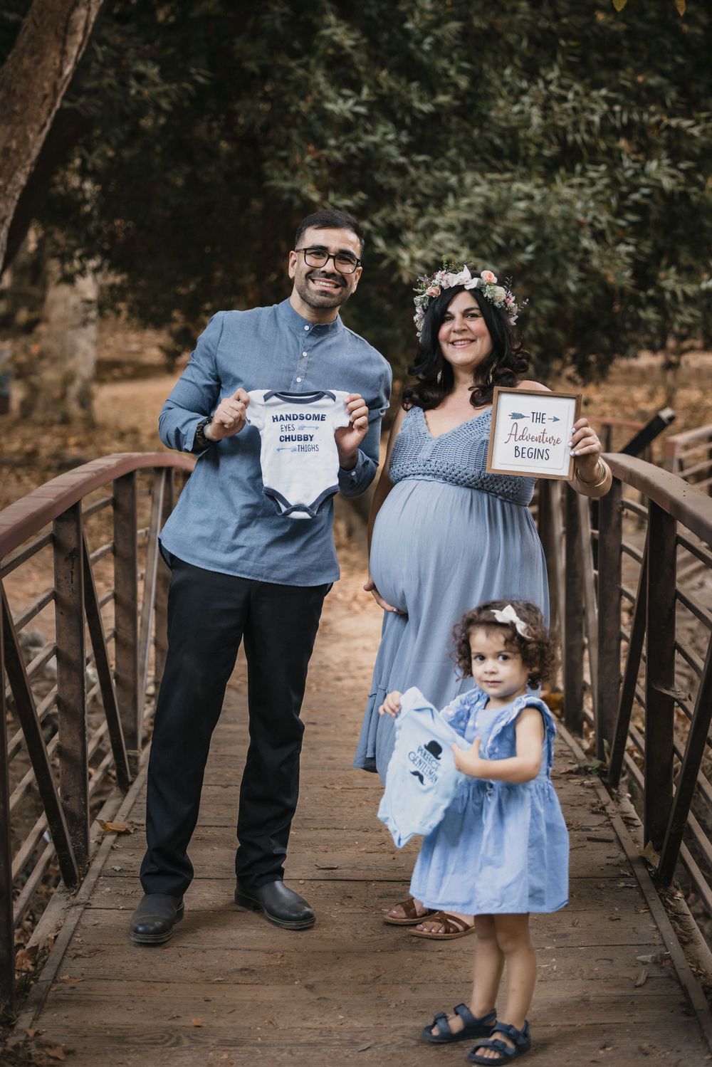 A family poses on a rustic bridge holding baby announcement signs while a toddler stands beside them.
