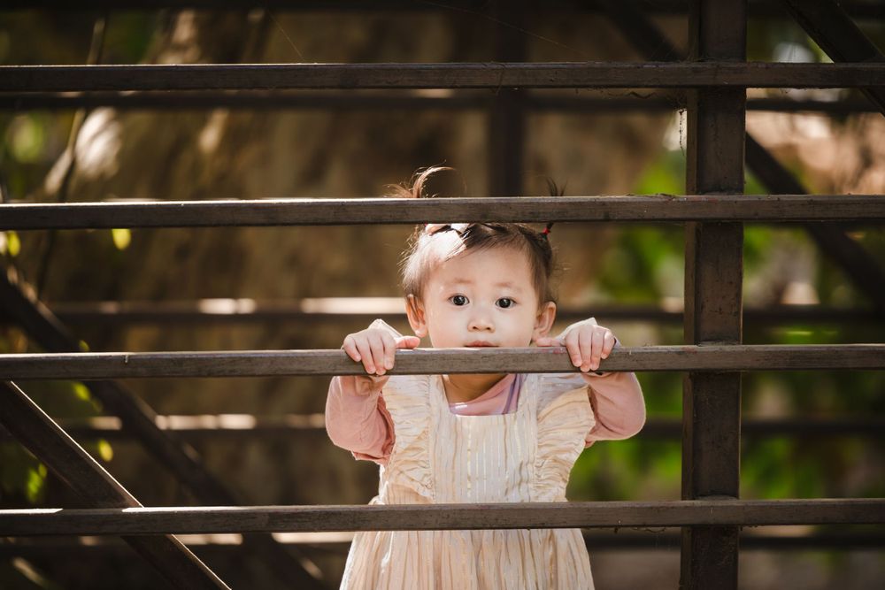 A toddler in a light dress peeks through metal railings while holding onto them outdoors.
