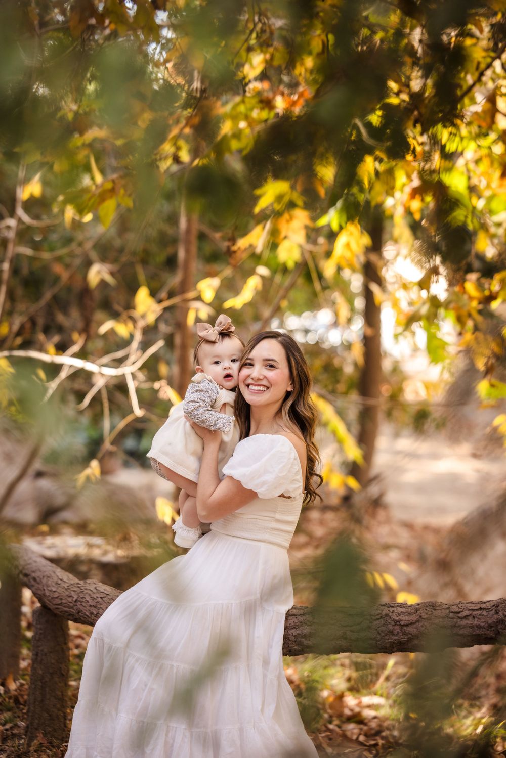 A white-dressed woman and baby share a joyful moment among golden autumn leaves in a natural outdoor setting.