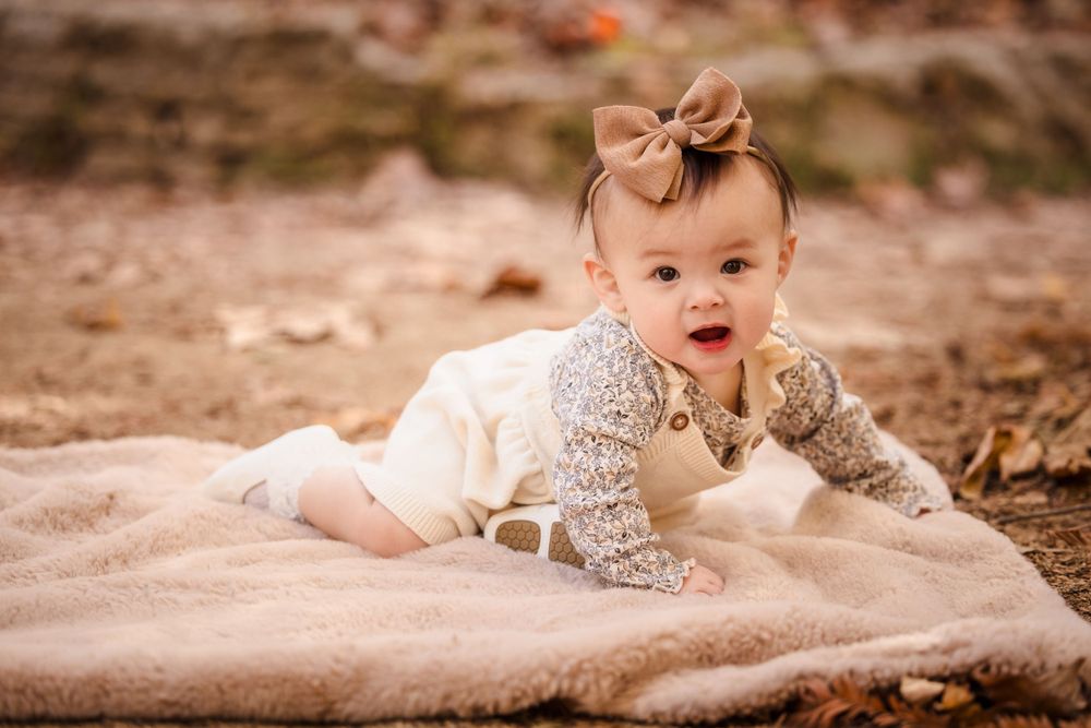 Baby wearing a bow crawls on a blanket surrounded by fallen leaves in an autumn setting.