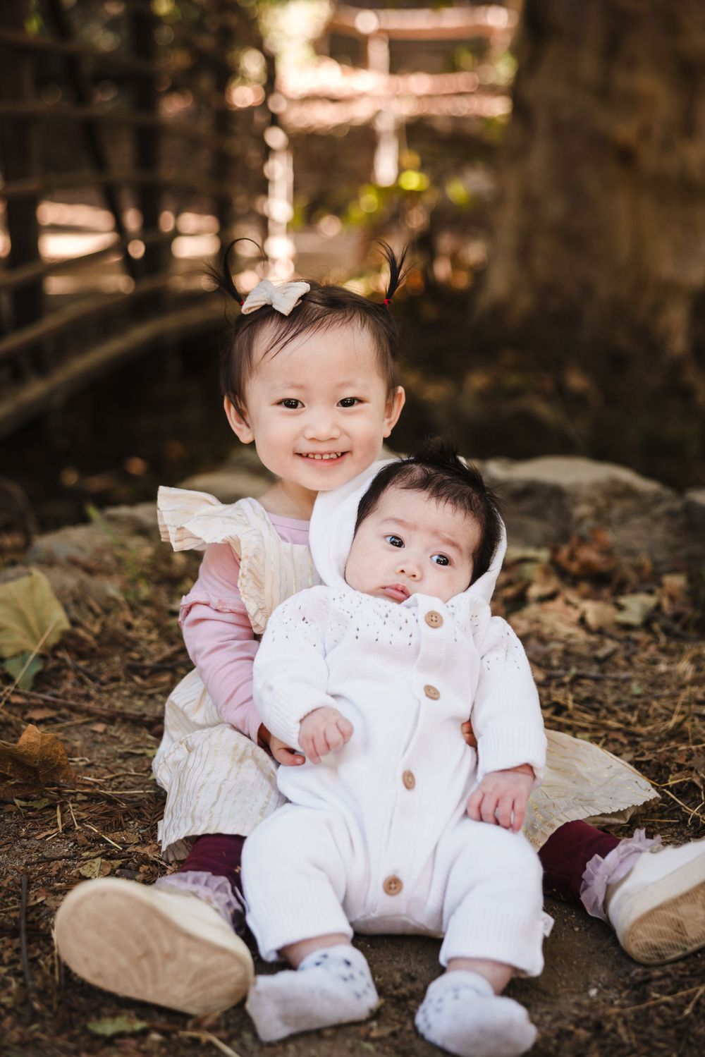 Two young children in white outfits sit together on autumn leaves in an outdoor setting.