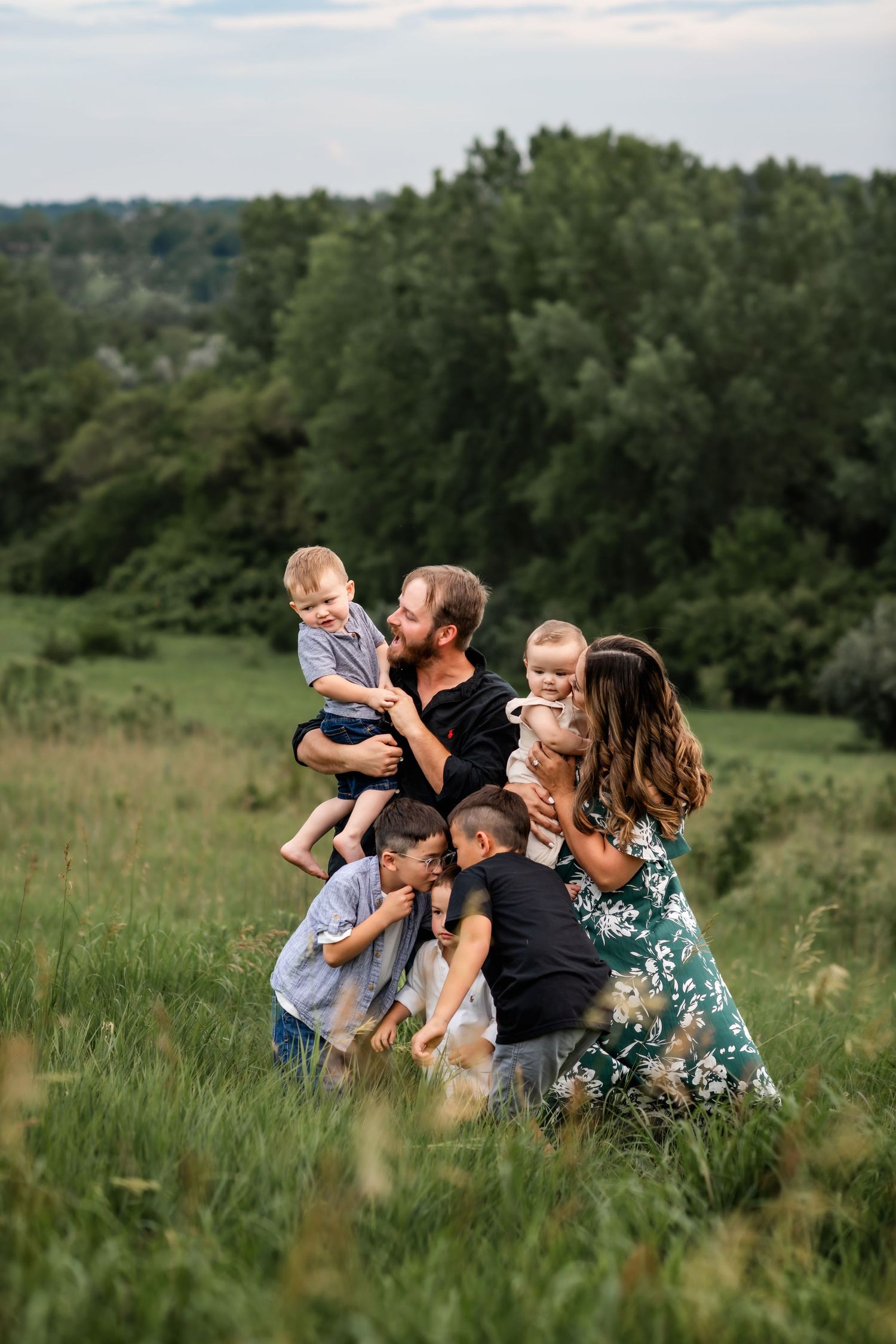 A family enjoys a candid moment together while kneeling in a green summer meadow with trees in the background.