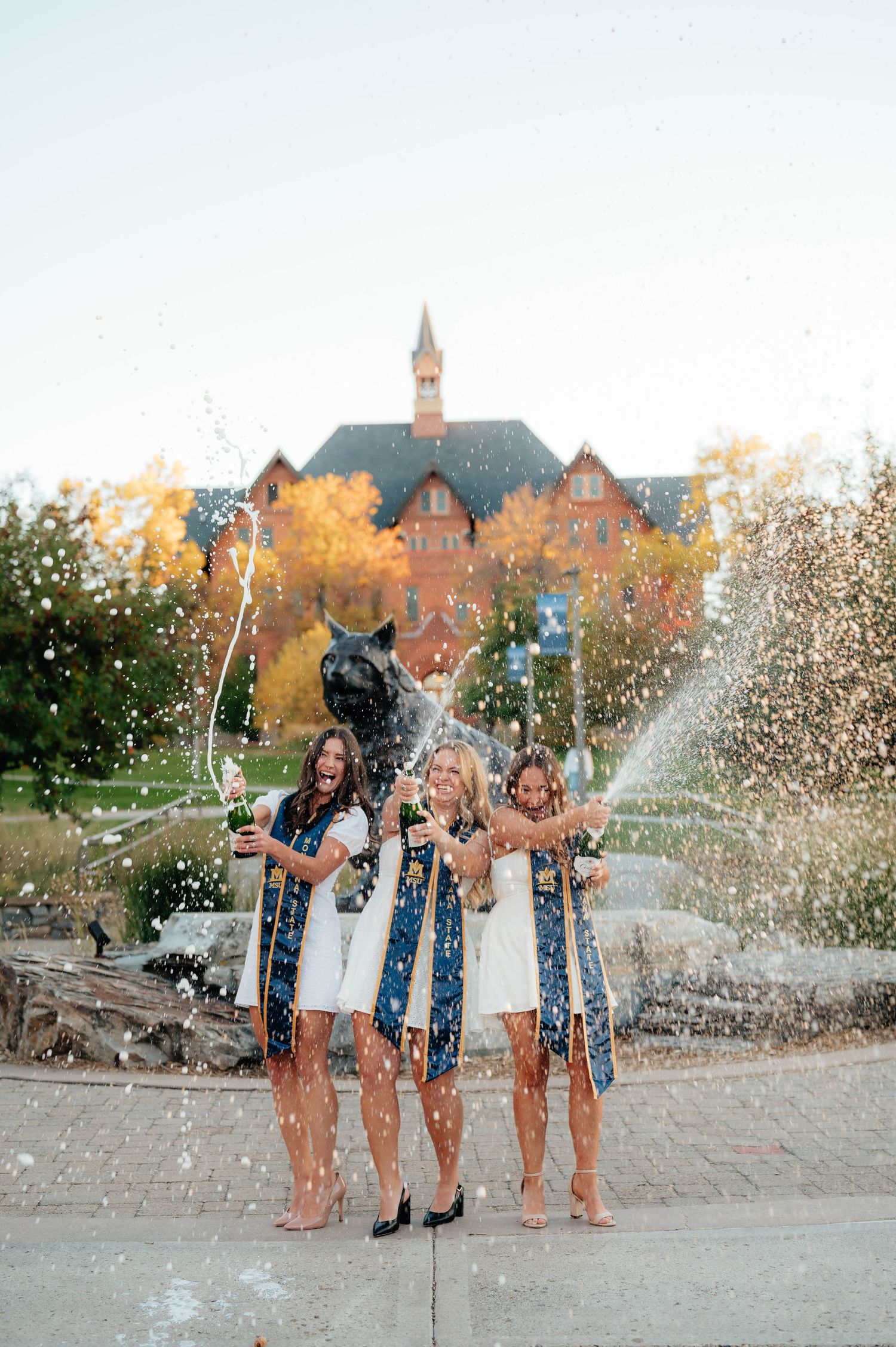 Three women in white dresses popping champagne near university bobcat statue with fall foliage backdrop.