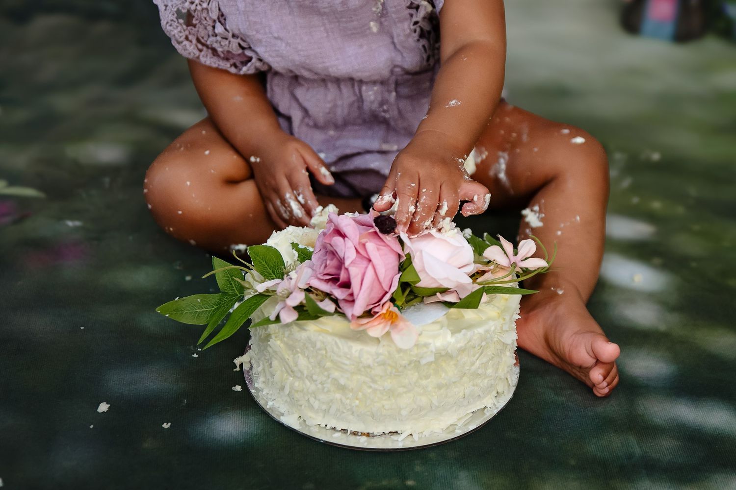 Small bare feet next to a white cake decorated with fresh pink and white flowers during an outdoor celebration. whk cakesmash photographer