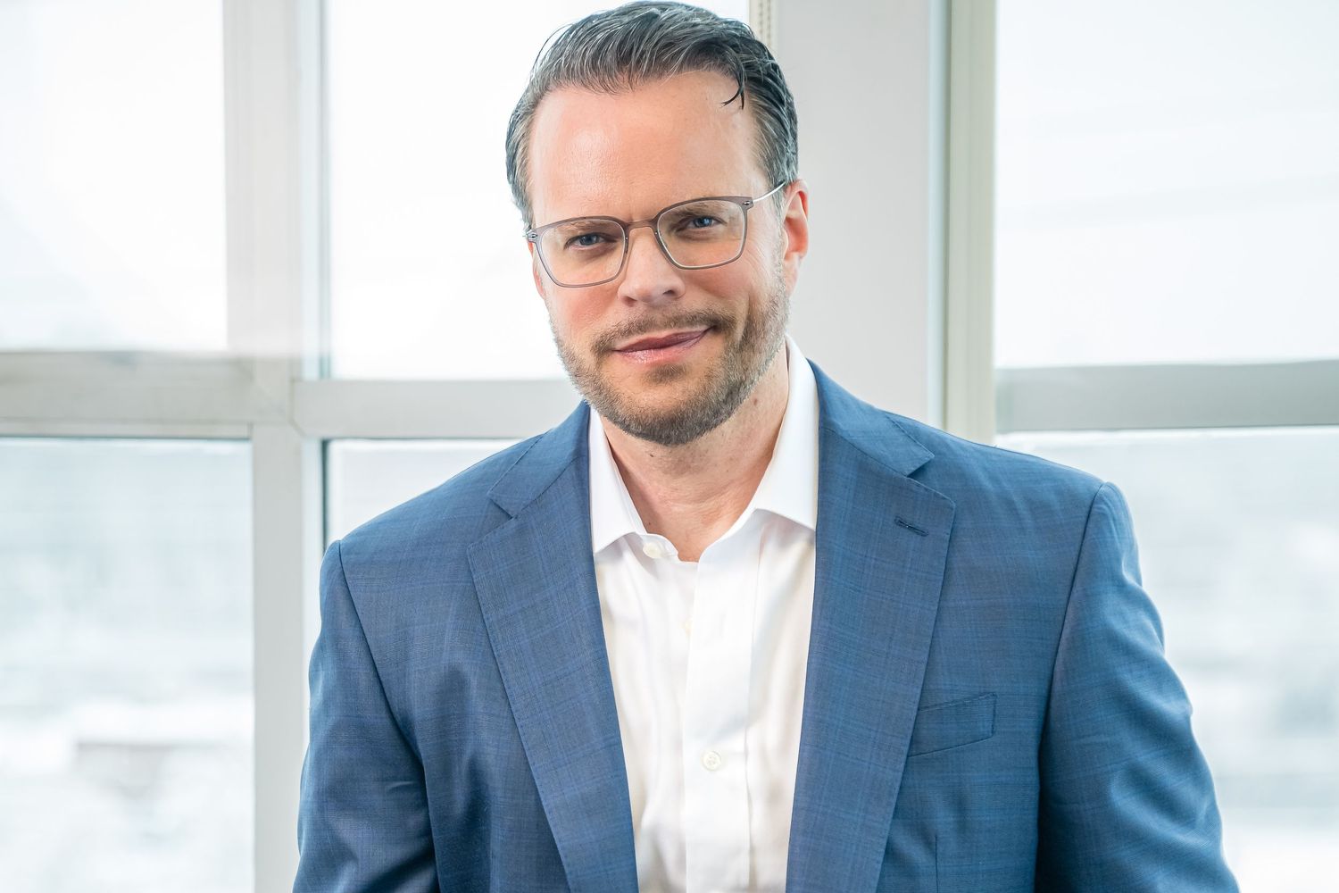 Professional wearing a blue suit jacket and glasses stands by a bright window in an office setting.