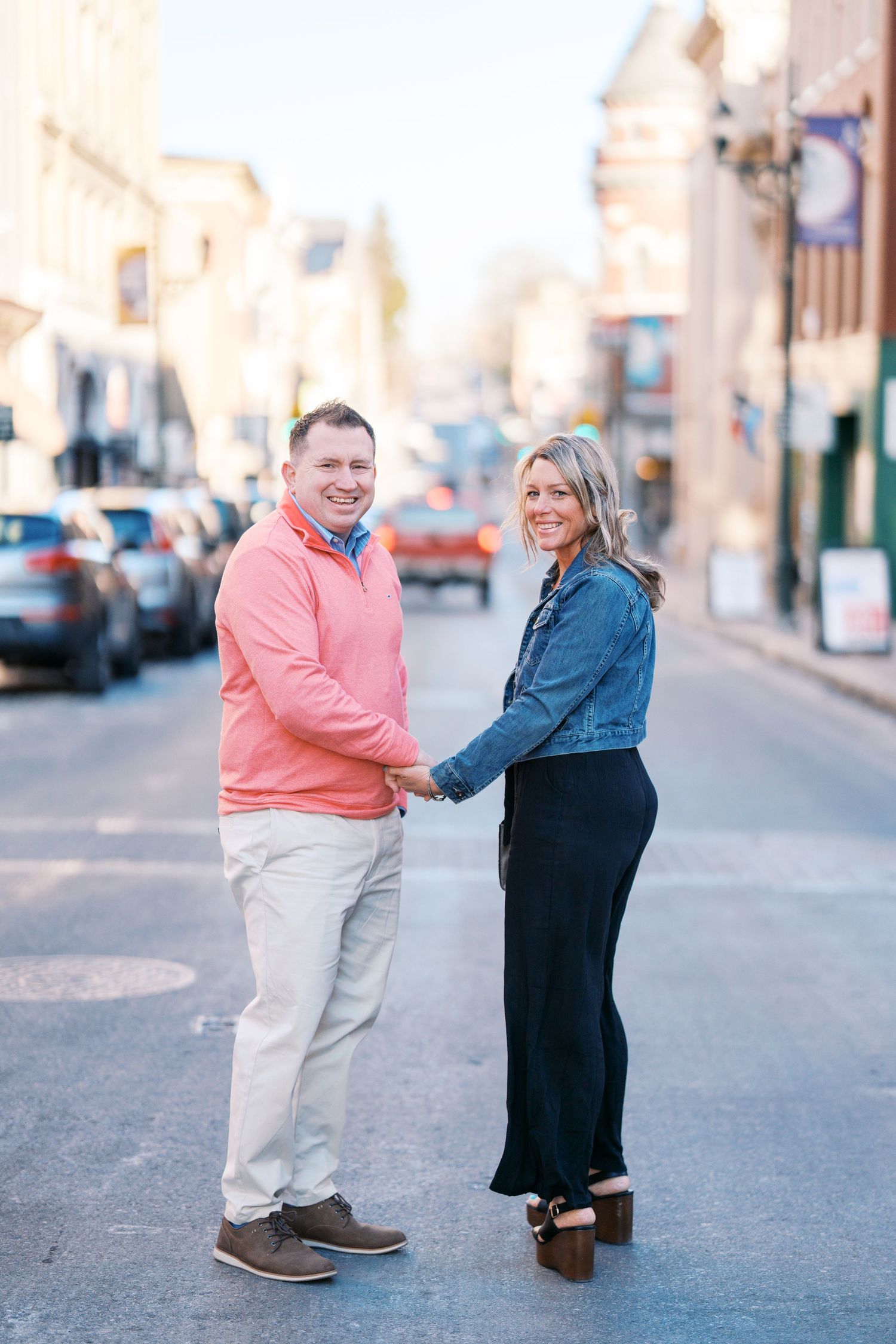 A couple holds hands while standing in the middle of a sunlit city street with buildings and parked cars in the background.