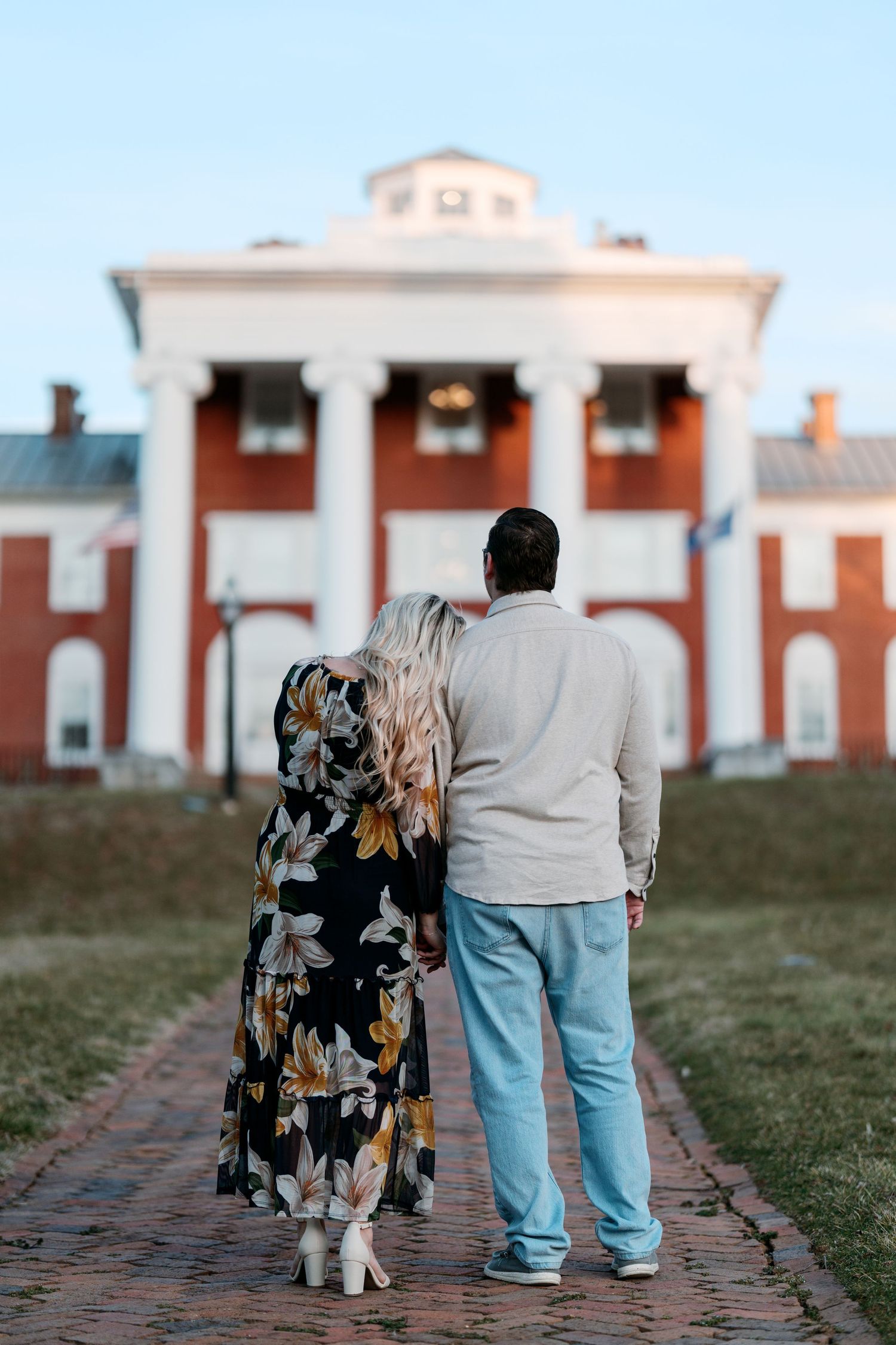 A couple stands on a brick path facing a grand red and white colonial mansion with pillars at sunset.