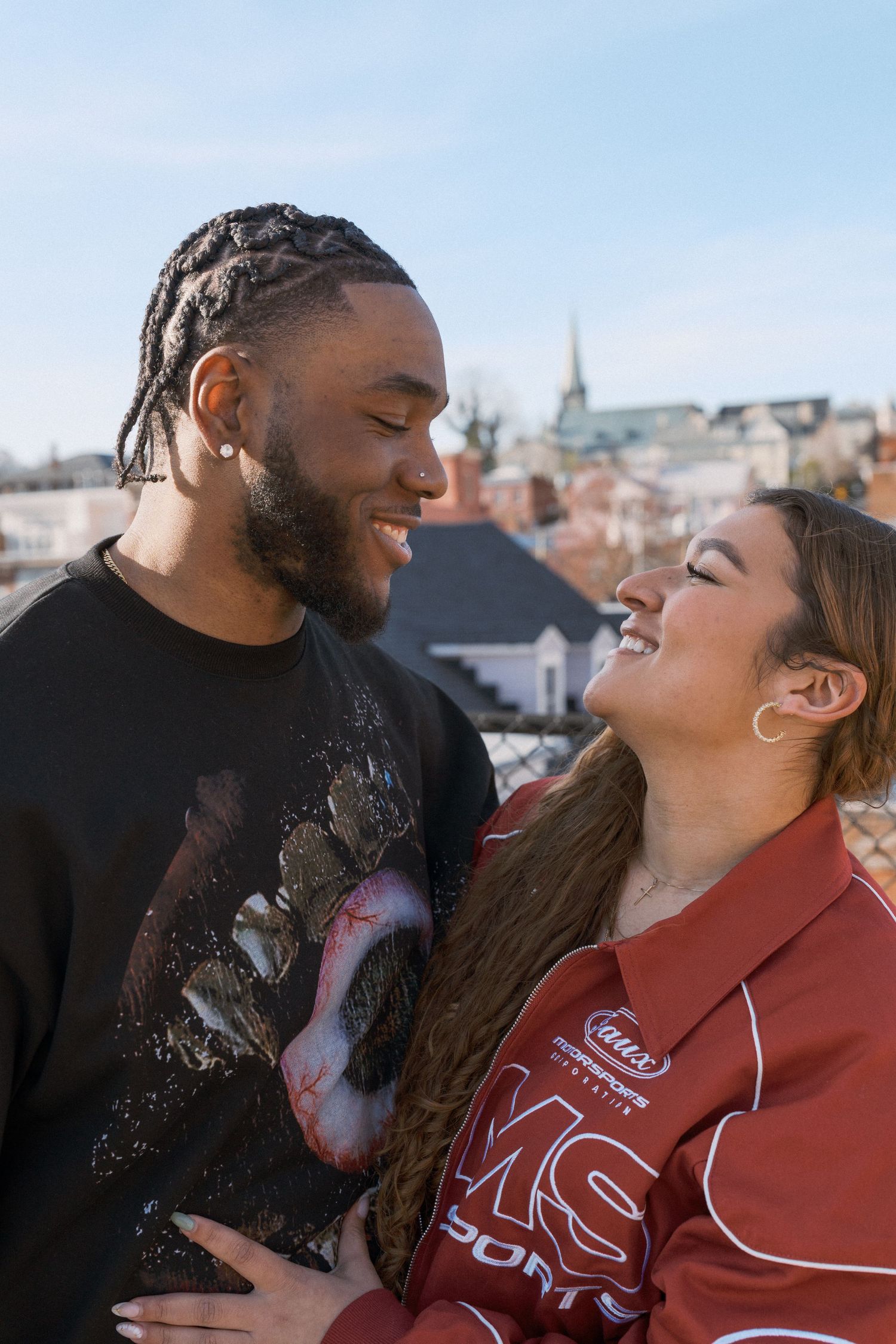 A happy young couple laughs together on a rooftop with a cityscape and church steeple visible in the background.