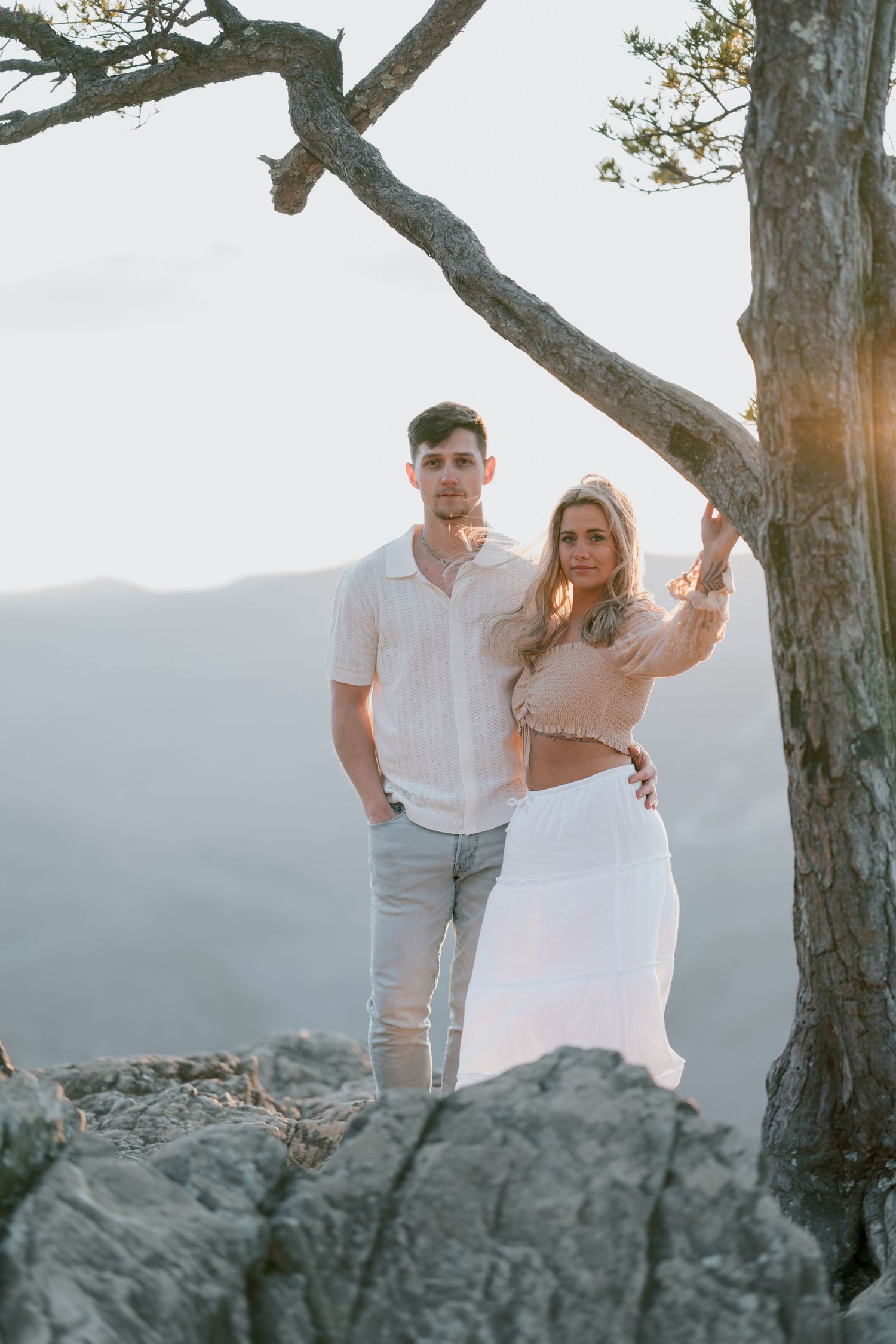 Two people pose romantically on a rocky cliff edge at sunset with a twisted tree branch above them.
