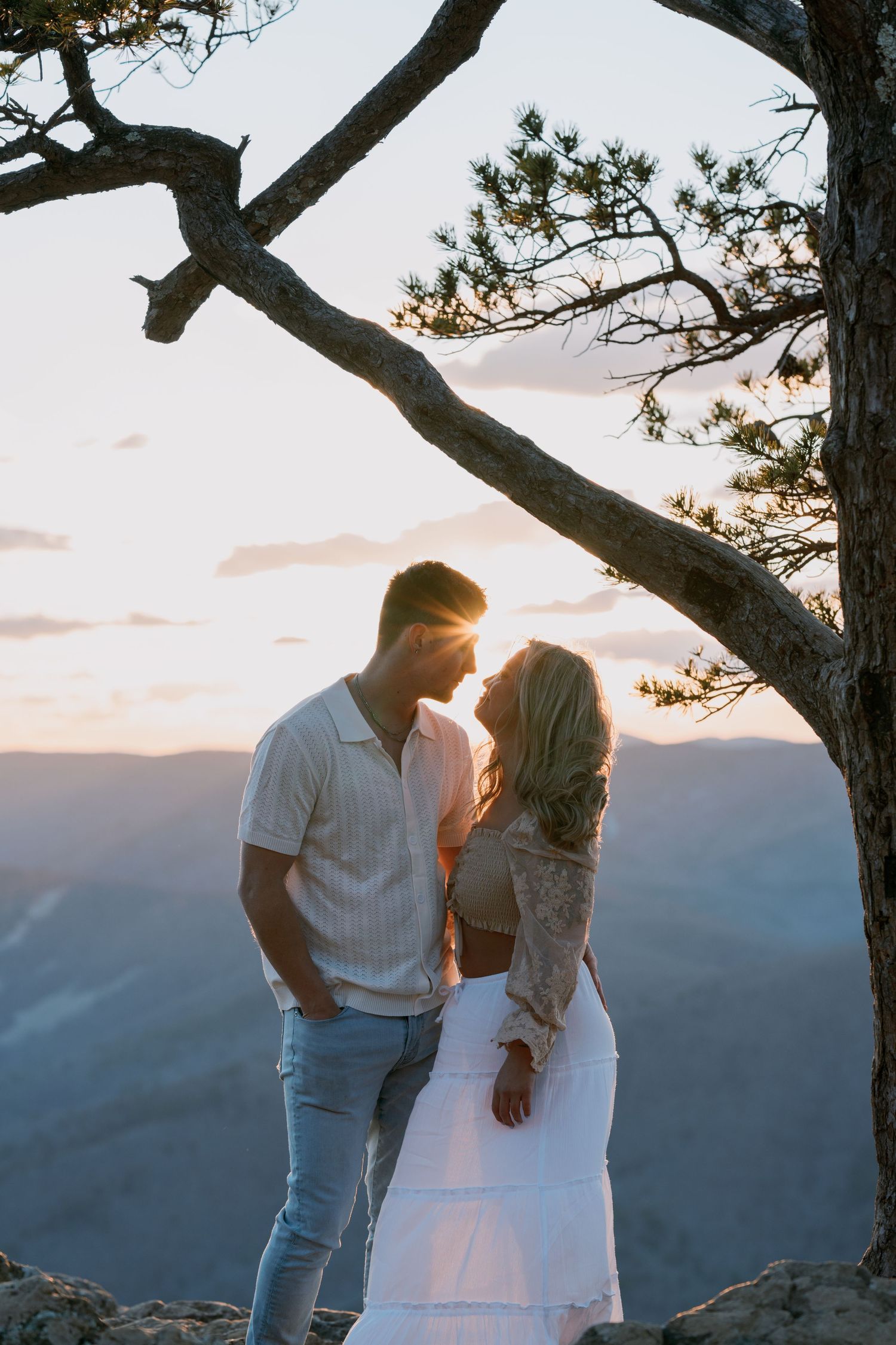 A romantic couple stands silhouetted against a mountain sunset, framed by a gnarled tree branch above.