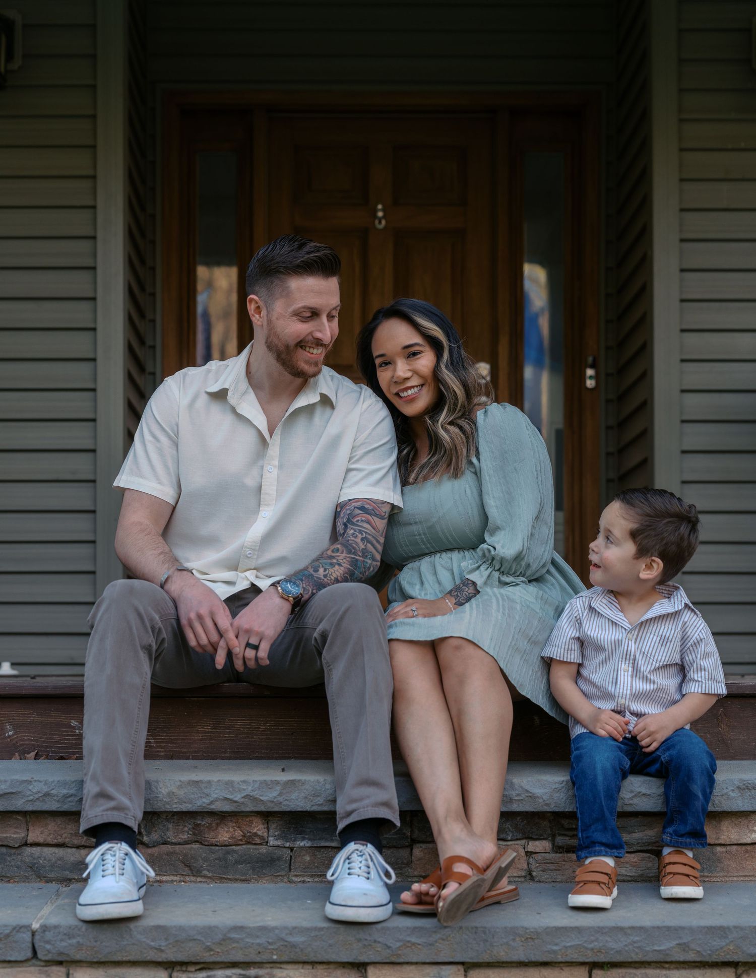 A happy family sits together on their front porch steps of a gray house on a sunny day.