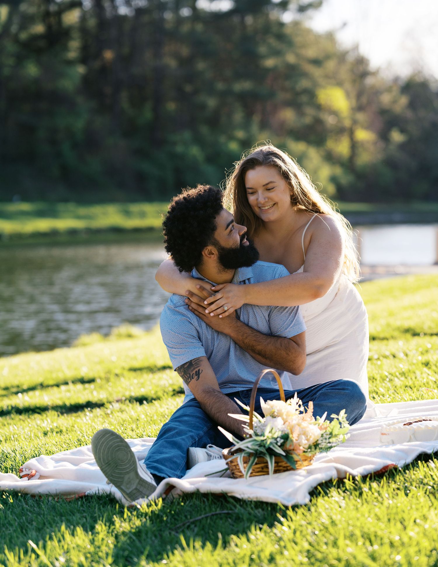 A couple enjoys a romantic picnic by a lake on a sunny day, sitting on a blanket with fresh flowers.