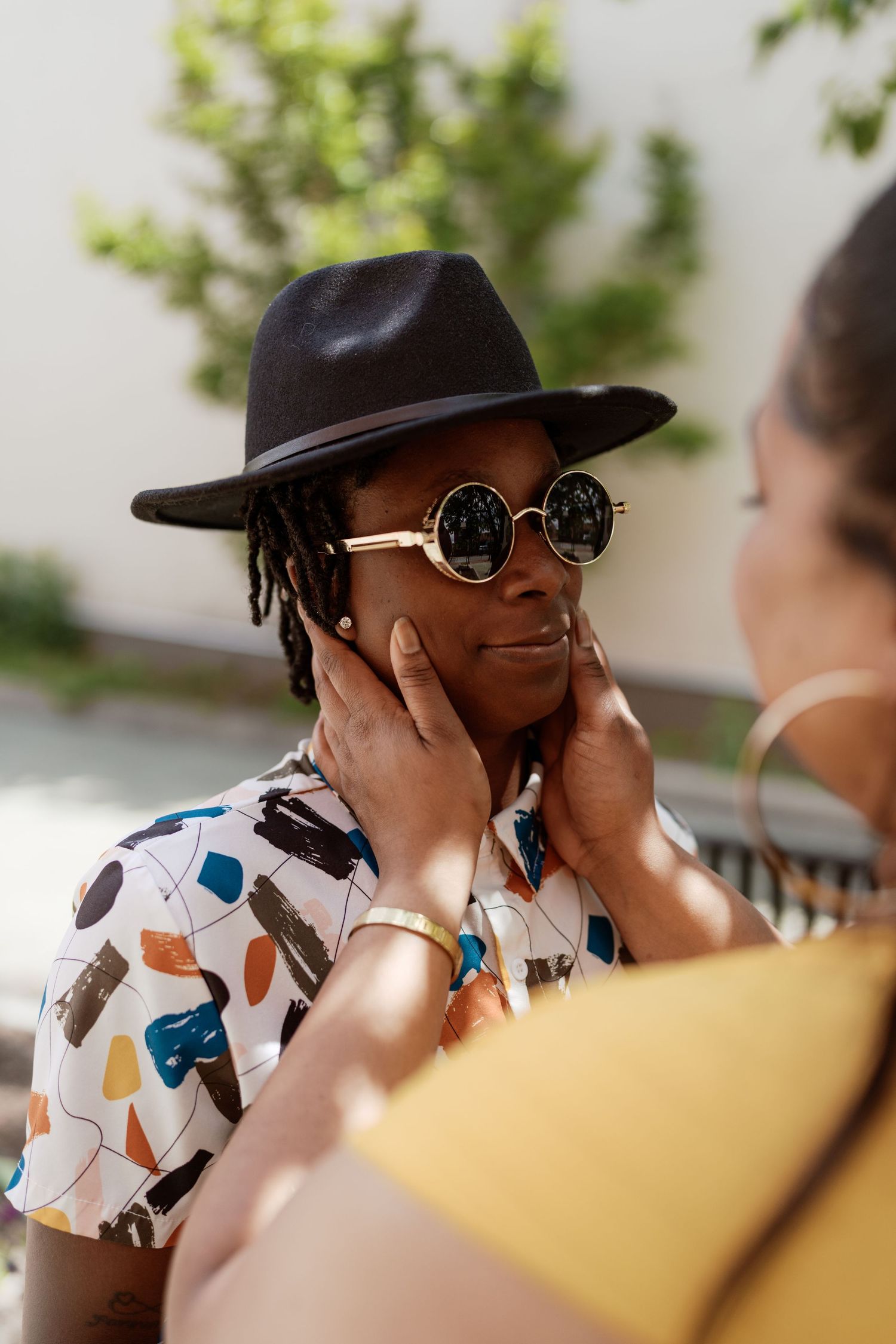 A stylish person wearing a fedora hat, round sunglasses and patterned shirt smiles while someone touches their face.