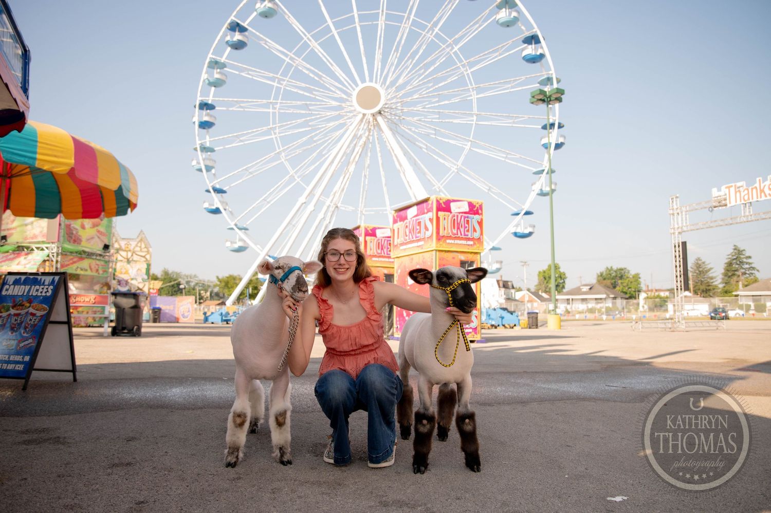 Woman sitting with two sheep in front of a colorful ferris wheel at a fair.