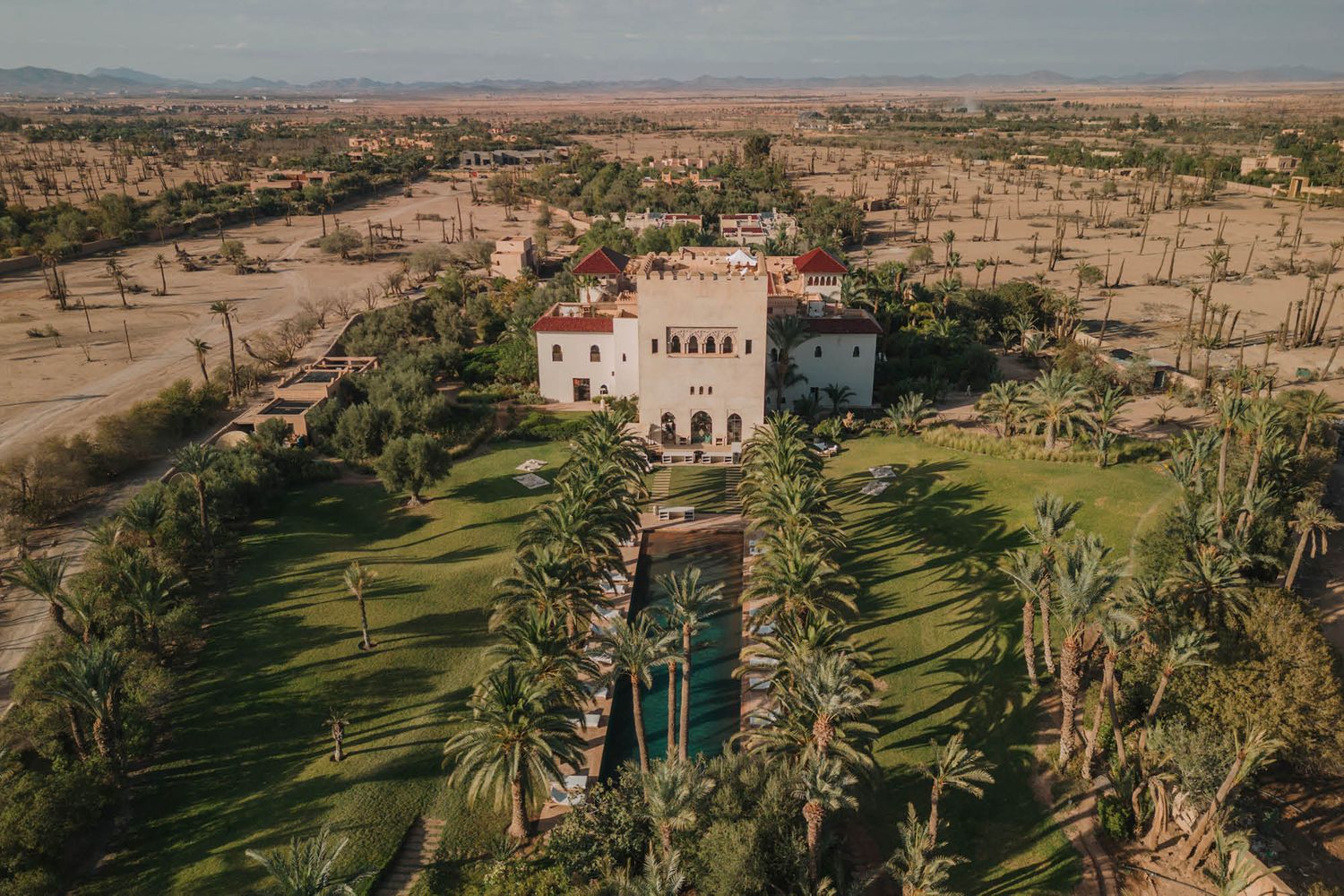 Luxury villa with manicured gardens and palm trees in a desert landscape viewed from above.