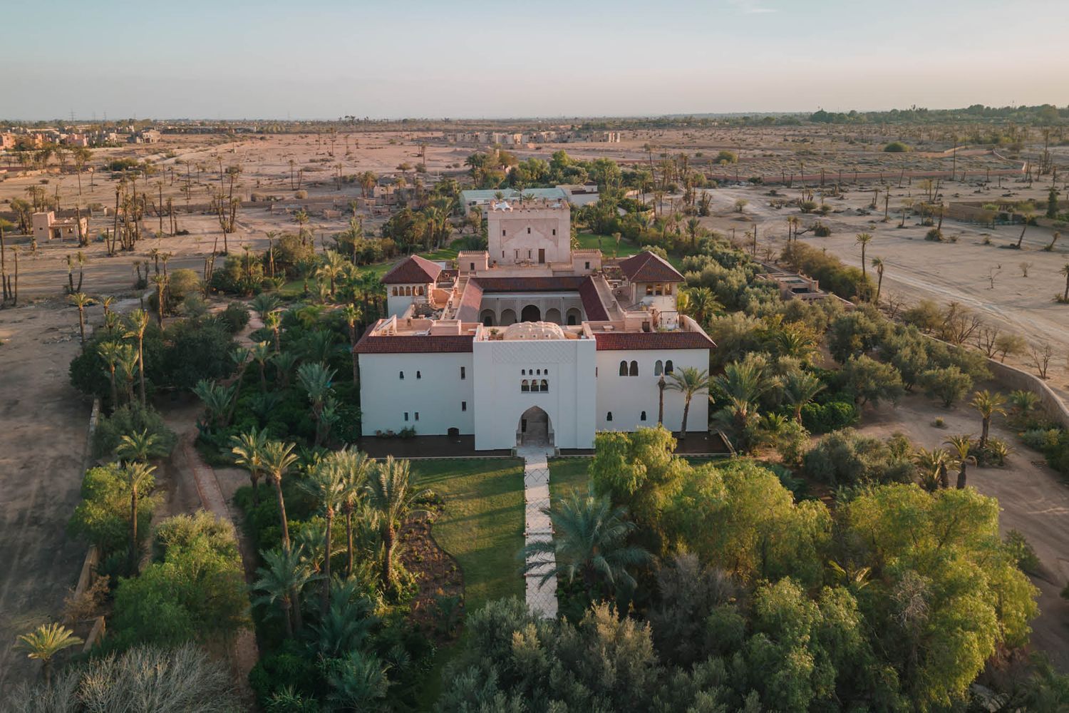 Aerial view of a white Spanish mission-style building surrounded by desert landscape and green trees at sunset.
