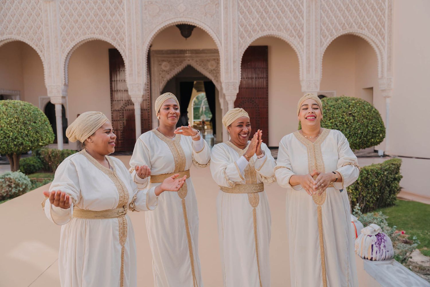 Group in flowing white gowns sharing joyful moments together in an ornate courtyard with arched walkways.