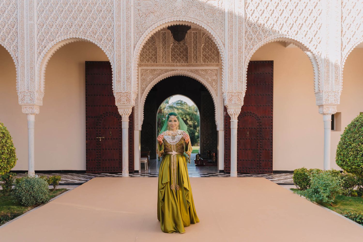 An elegant figure in a flowing gold dress poses beneath ornate Moroccan-style archways and architectural details.