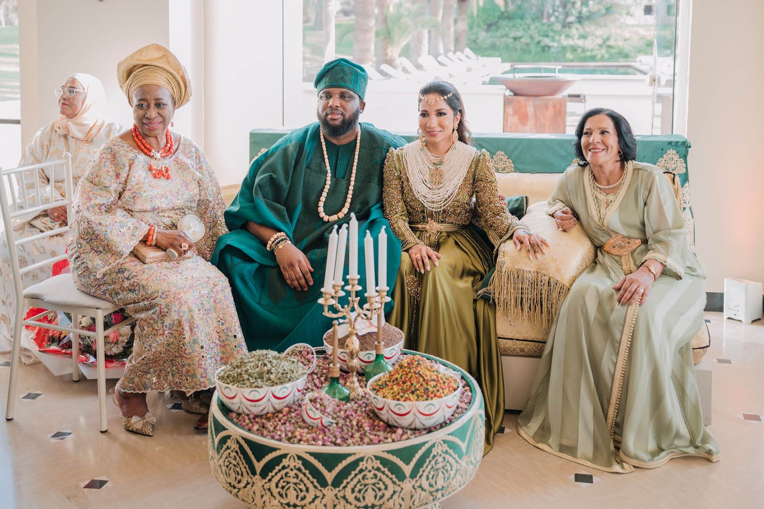 A wedding celebration with guests in traditional African formal wear seated around an ornate table with refreshments.
