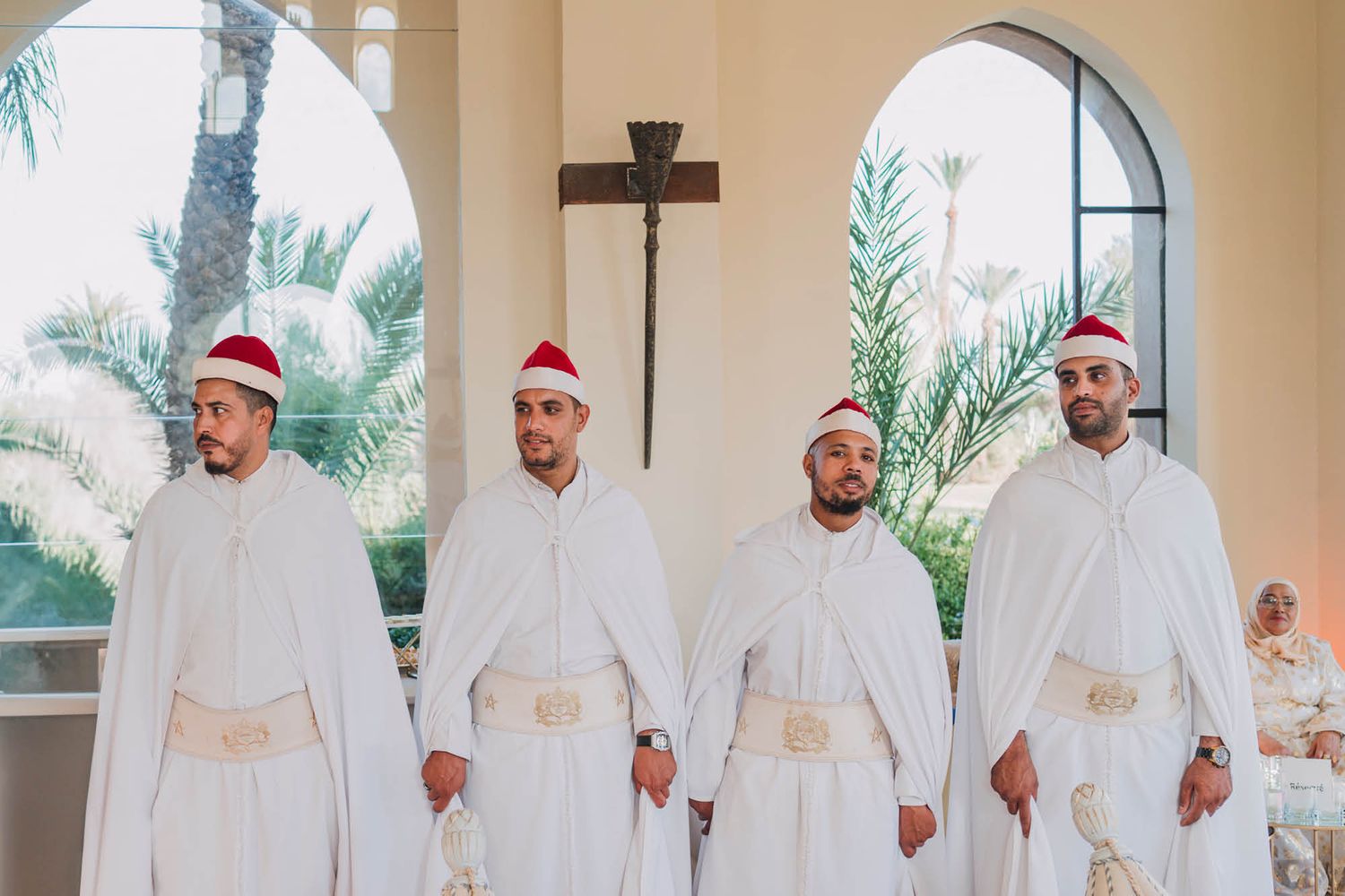 Four people in white robes and red Santa hats stand together inside a chapel with palm trees visible through arched windows.