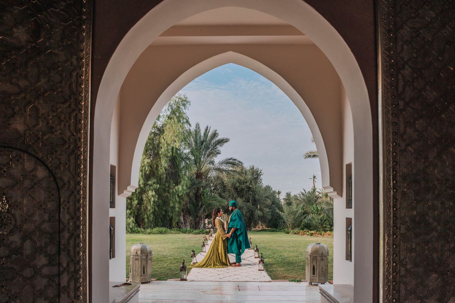 View through ornate archway showing couple in traditional attire walking together in palace gardens.
