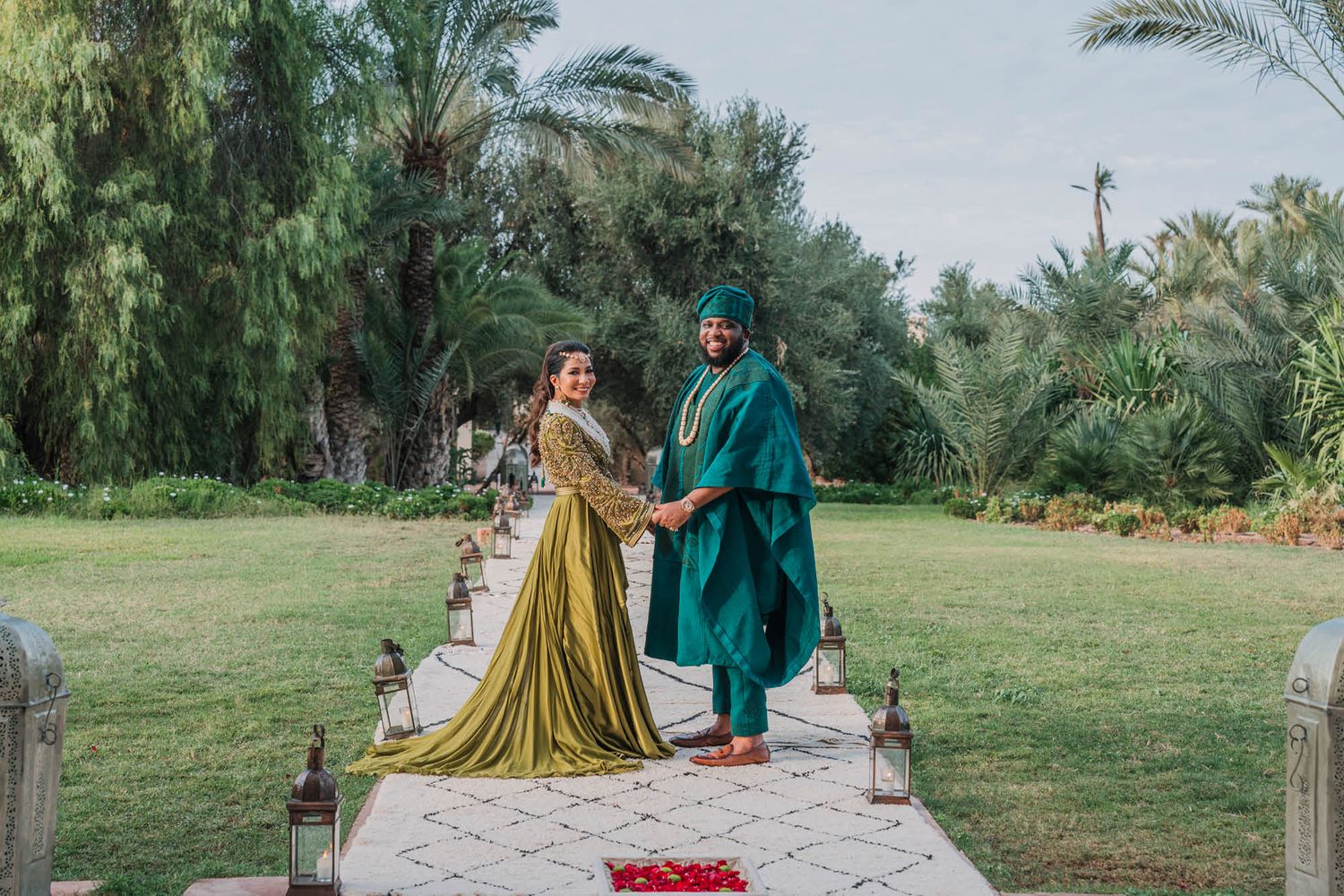 A couple in elegant attire stands on a path lined with lanterns amidst a lush tropical garden setting during a ceremony.