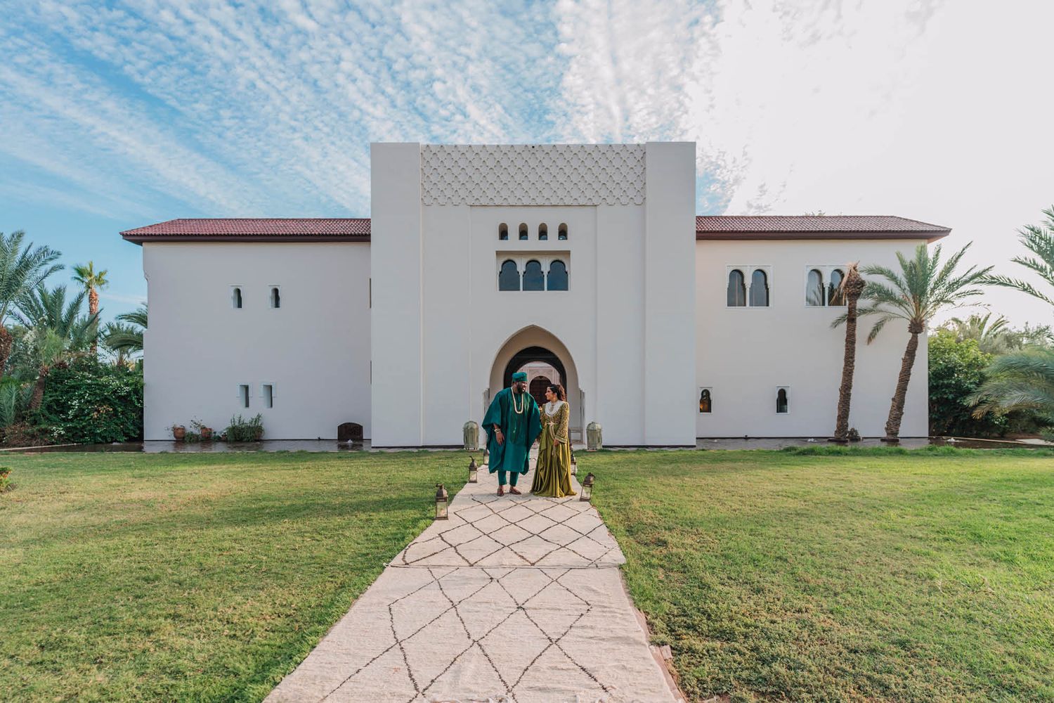 Two people in coordinated teal outfits stand before a white Moorish-style building with arched doorways and palm trees.