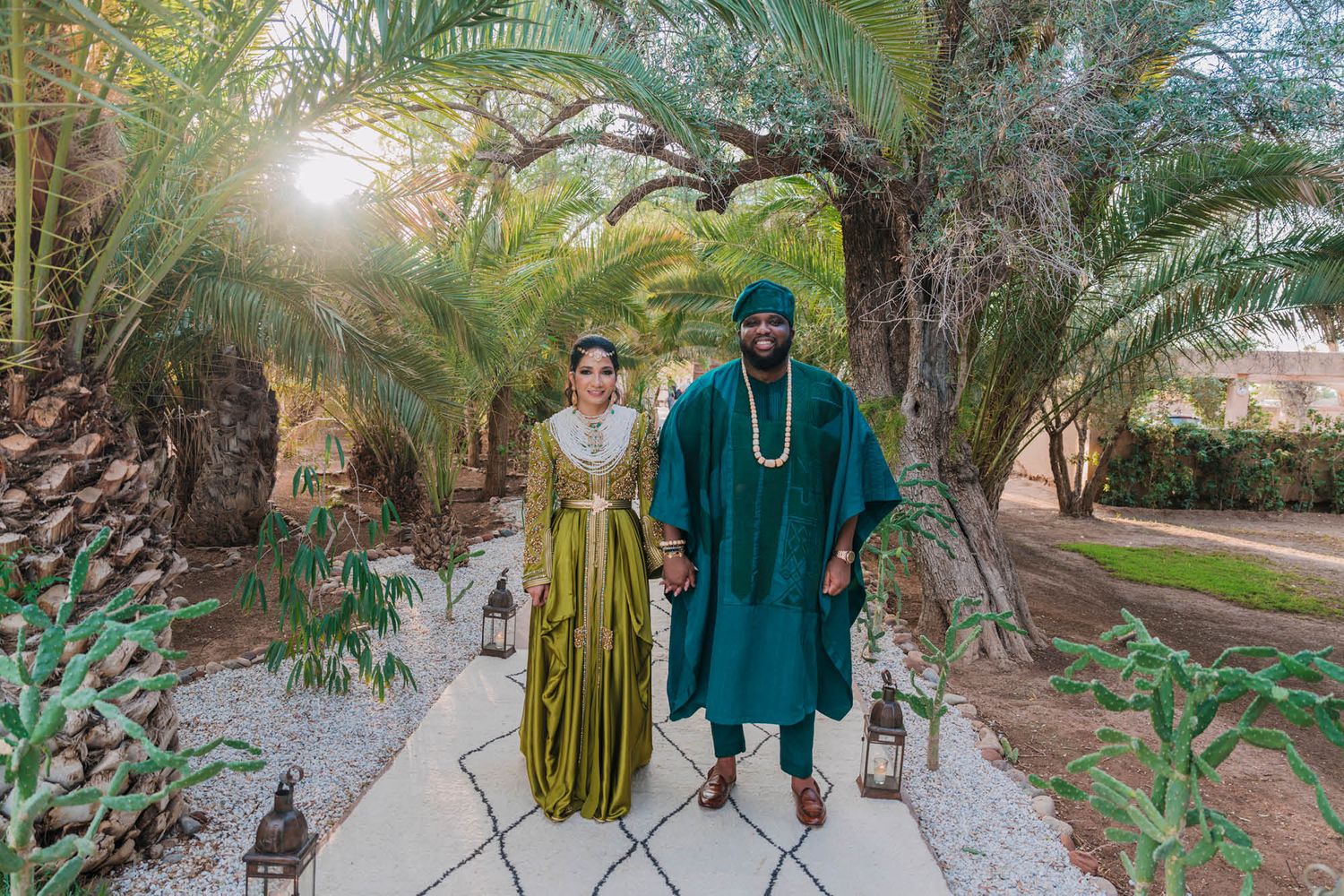 A couple in traditional African attire walks along a palm-lined garden path carrying ornate lanterns at sunset.