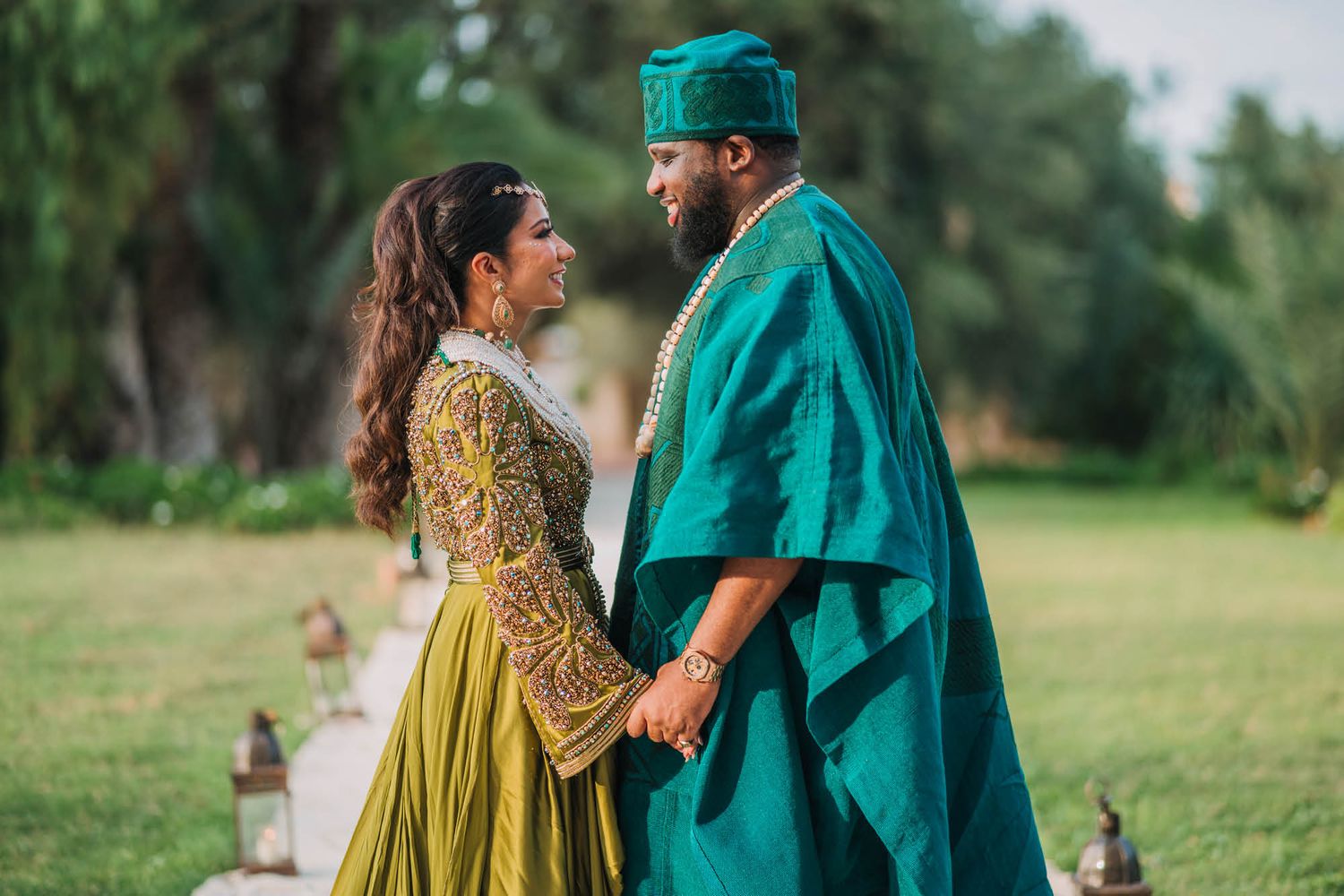 A couple in elegant green and gold traditional wedding attire holding hands outdoors in a garden setting.