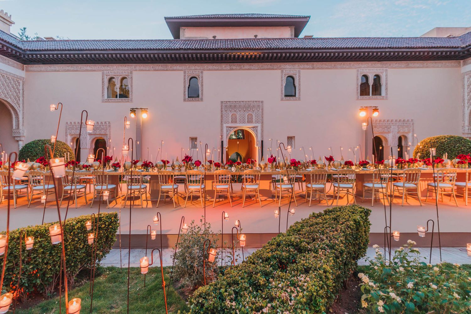 Elegant outdoor wedding reception setup with orange chairs and candles at a Moroccan-style venue at sunset.