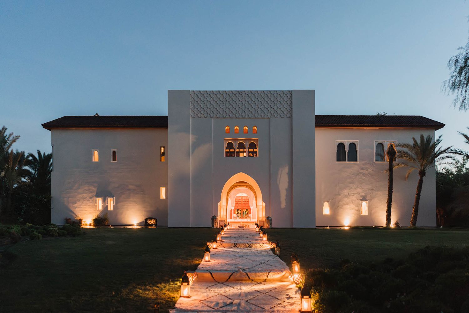 A white Mediterranean-style villa illuminated at night with a lit fountain path leading to an arched entrance.