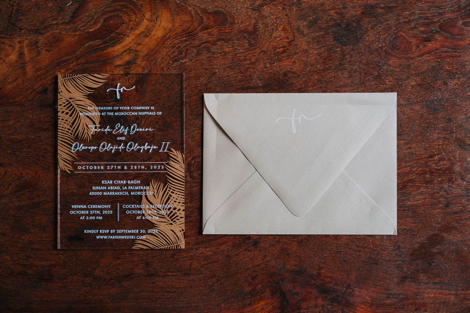 A wooden invitation with gold leaf details and white envelope on a dark wooden surface.