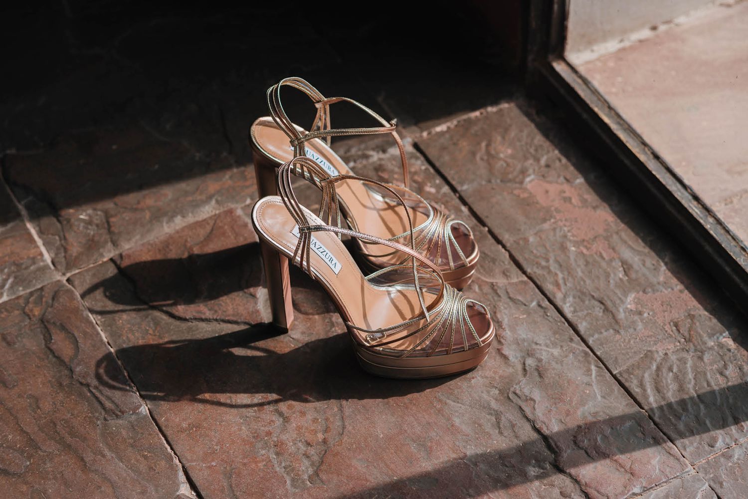 Elegant metallic strappy wedding heels sitting on stone tiles in natural lighting.
