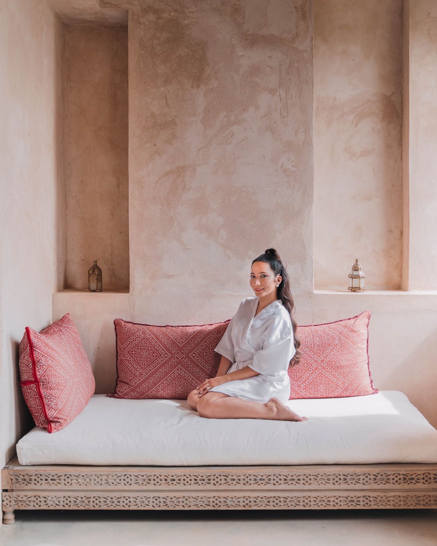 A comfortable daybed with pink cushions and traditional carved base sits against a textured beige wall with an alcove niche.