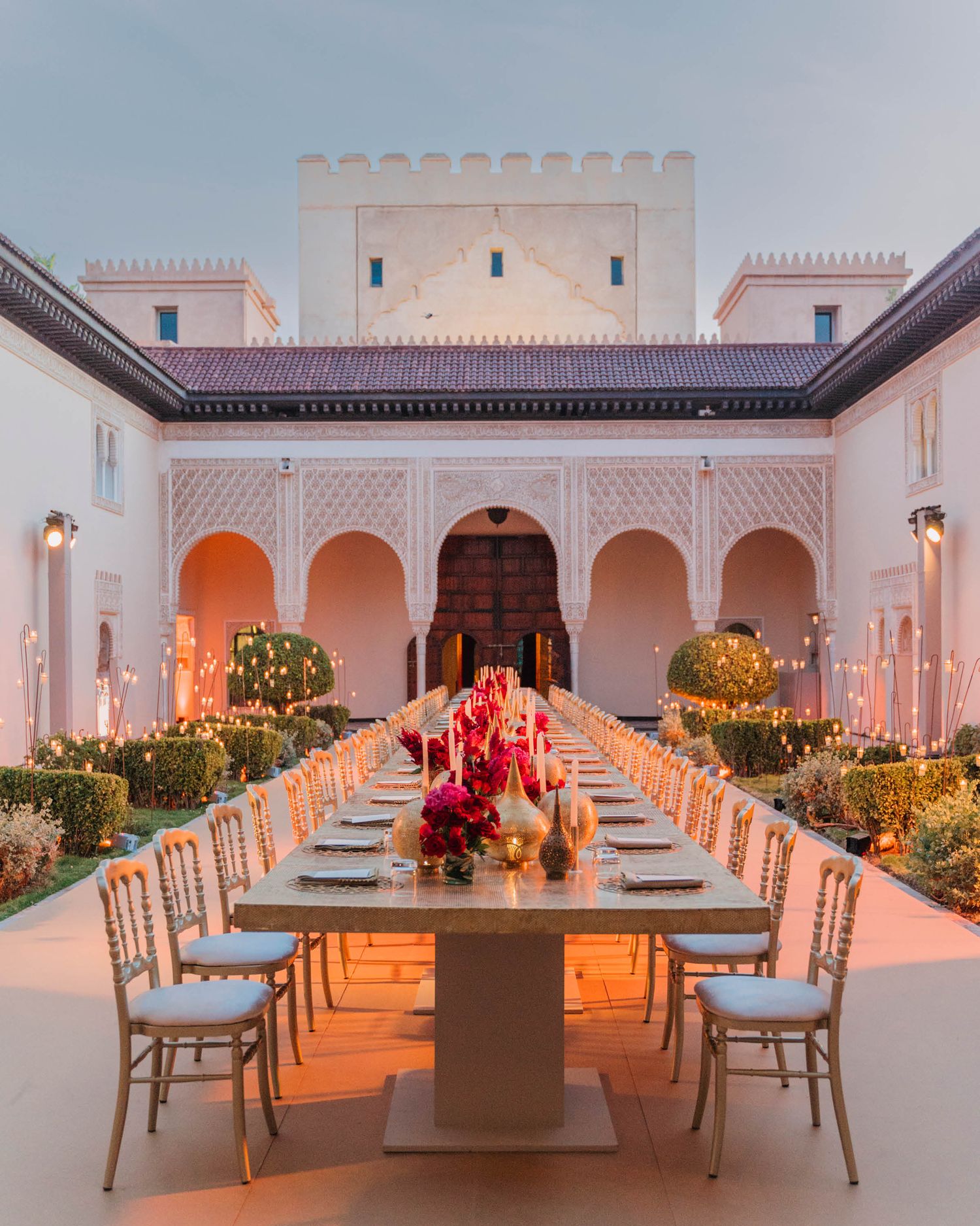 Elegant outdoor dining setup with long table and white chairs under Moroccan-style white arches illuminated at dusk.