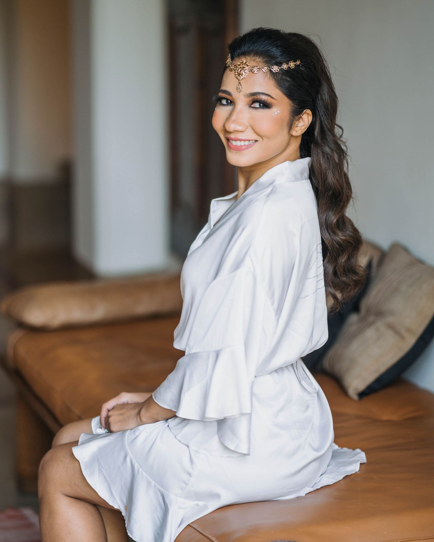 Someone wearing a white satin robe smiles while sitting on a brown couch, adorned with sparkly hair accessories.