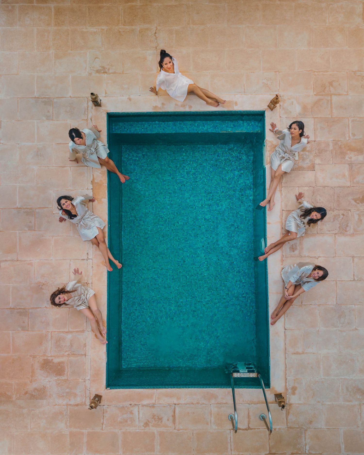 An aerial view of a swimming pool bordered by people lying on its edges in a symmetrical formation against beige stone walls.