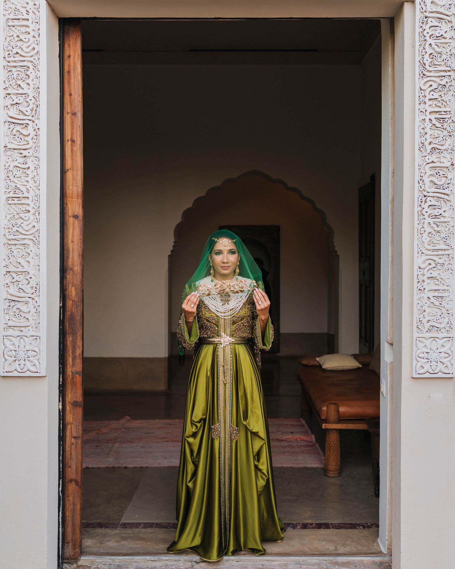 A person wearing a traditional green and gold silk gown with a matching headscarf stands in an ornate doorway.