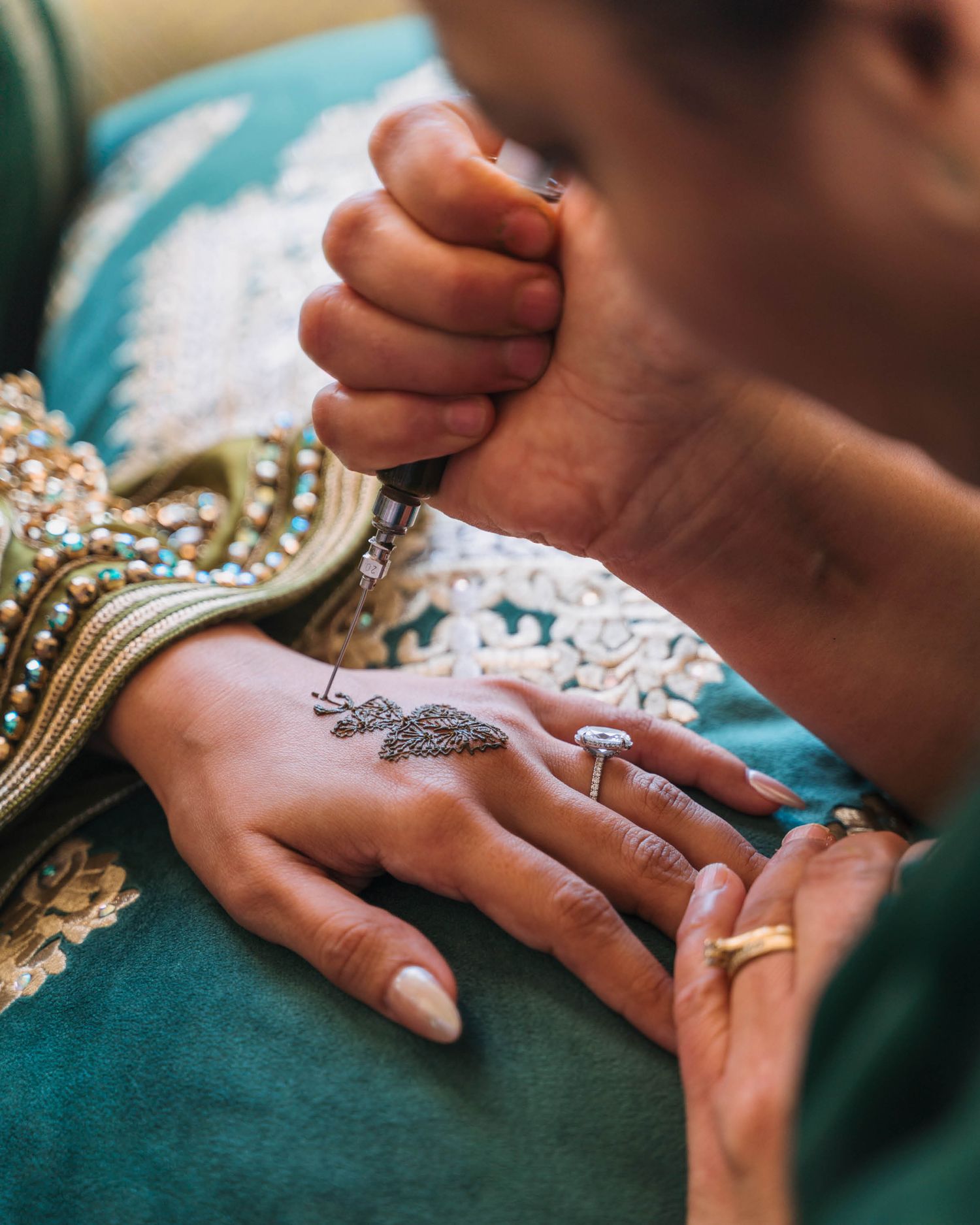 Close-up of hands applying intricate henna design on palm with jewelry against turquoise fabric background.