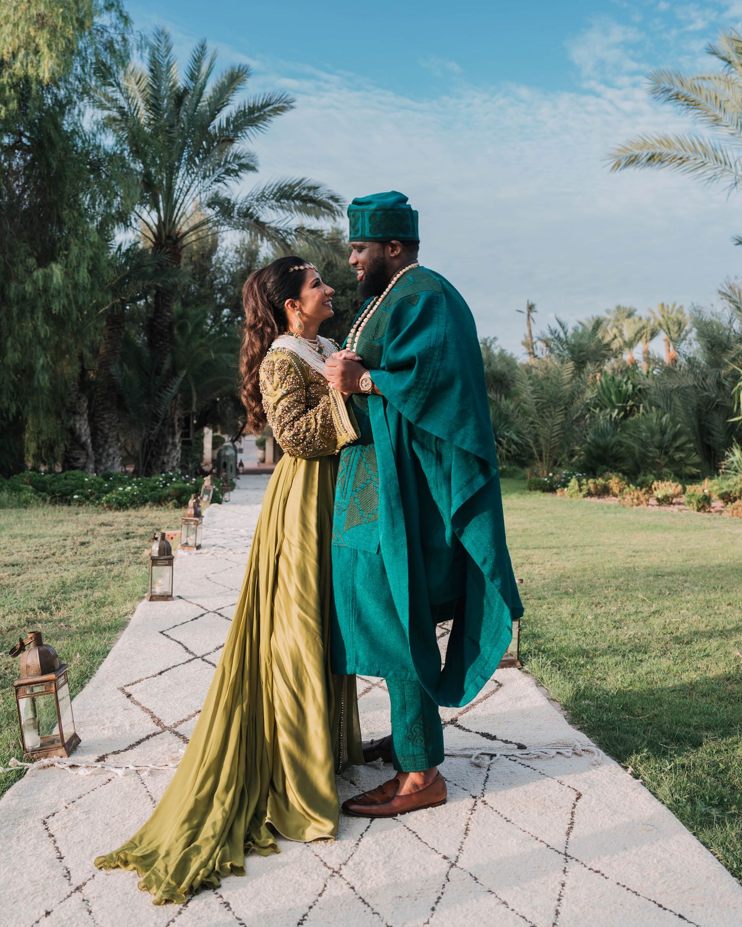 A couple in traditional attire, with a green robe and yellow dress, stand together on a stone path surrounded by palm trees.