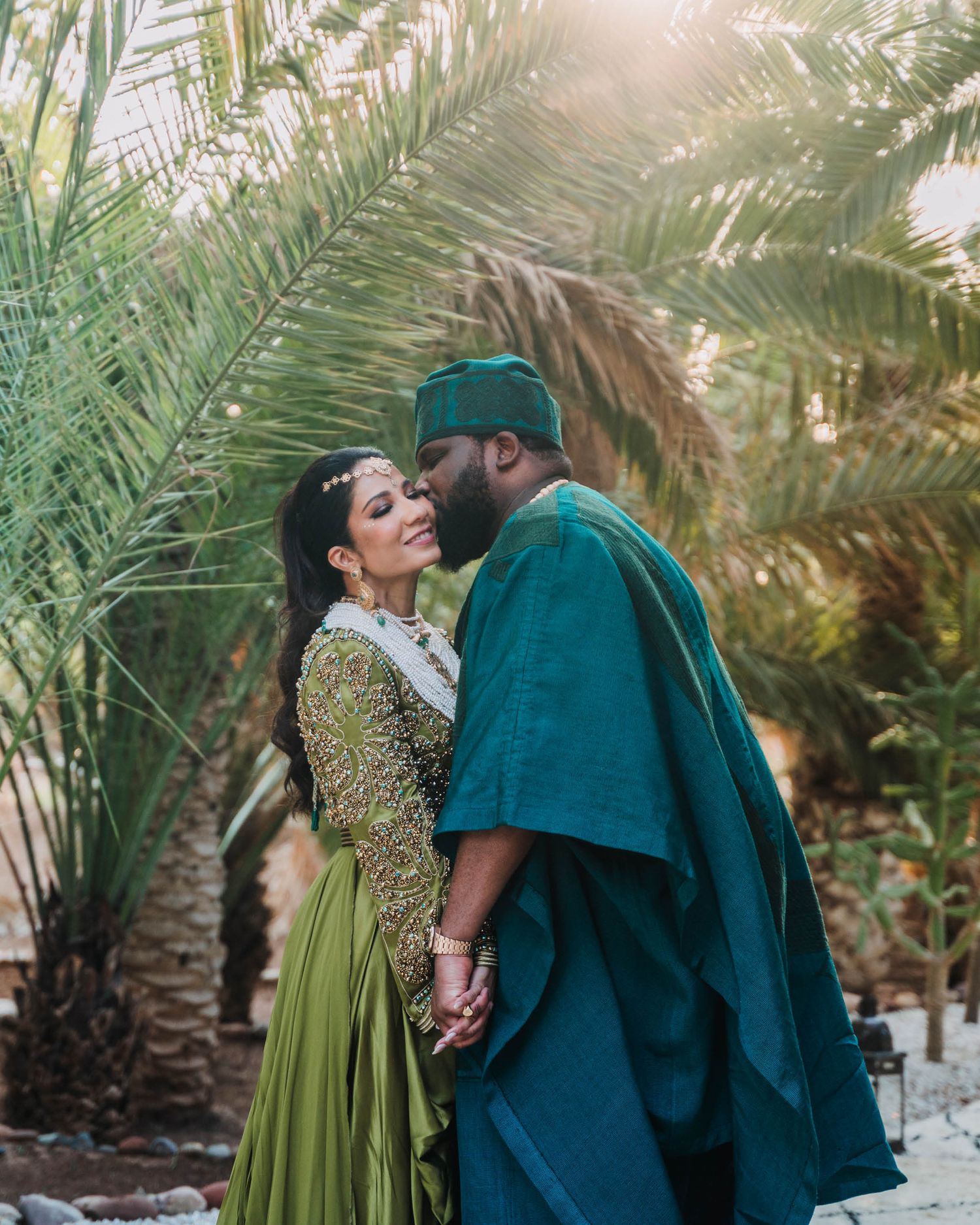 A romantic couple embraces among palm trees wearing elegant traditional outfits in green and teal tones.