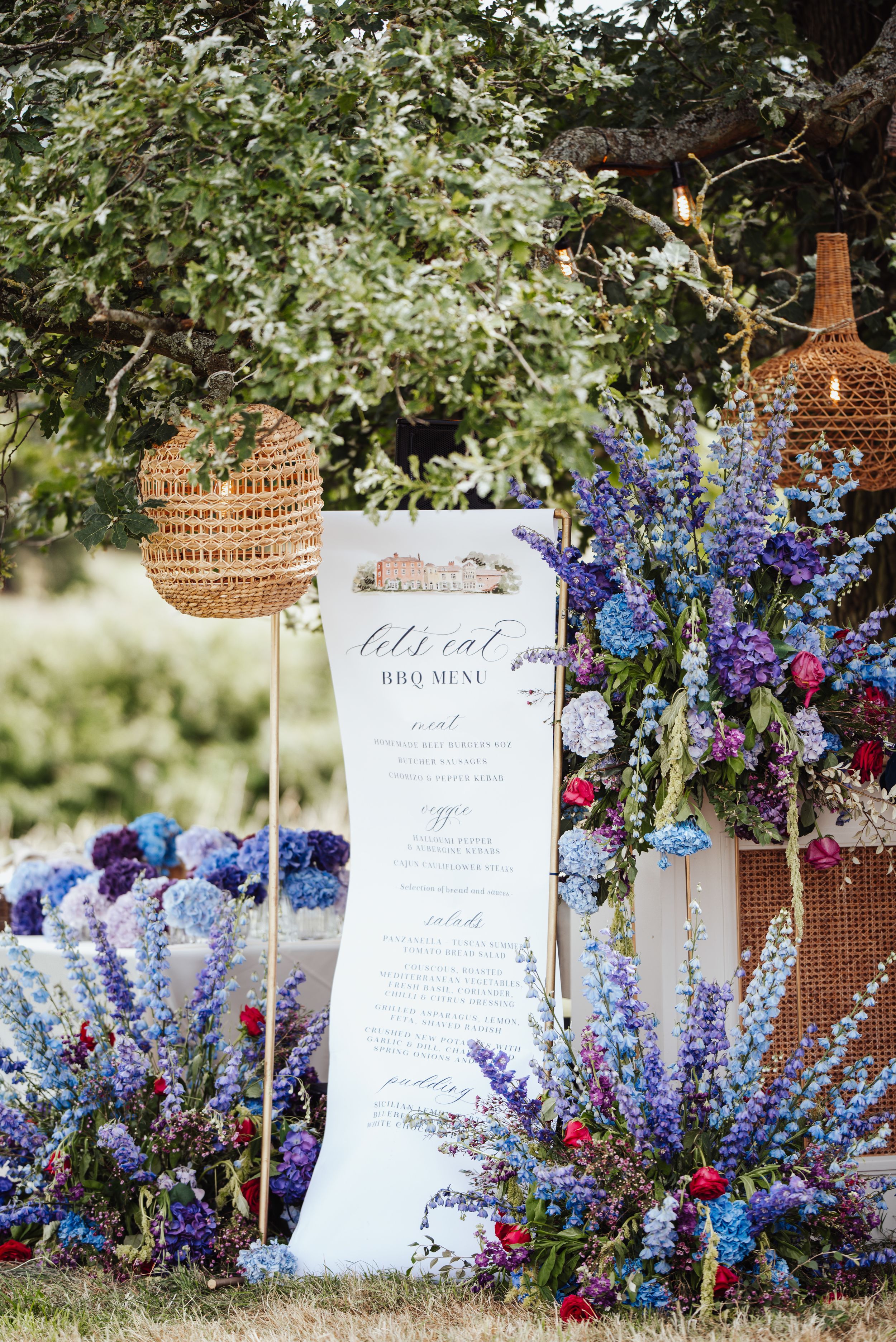 Elegant outdoor wedding menu display with rattan lanterns and vibrant blue delphinium and wildflower arrangements.